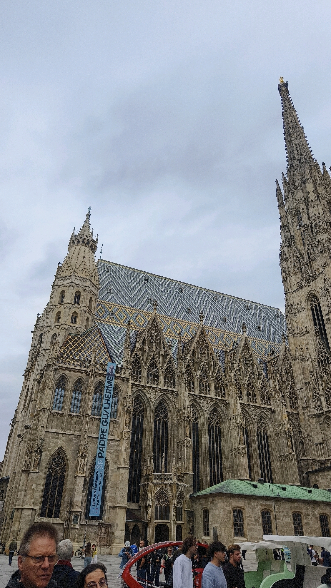 The image captures a wide-angle view of St. Stephen's Cathedral in Vienna, Austria, under a cloudy sky. The cathedral's intricate Gothic architecture is prominently displayed, with its distinctive patterned roof featuring zigzags of gray and yellow tiles. A tall, spired tower with a golden statue at its peak rises to the right of the main structure. On the left side of the cathedral, a smaller tower is visible. A blue banner with German text, reading "PADRE GU/LHERME", hangs vertically down the facade. In the foreground, several people are gathered. Two individuals are in focus in the lower left corner, with one man wearing glasses and the other a woman with dark hair. A red open-air vehicle, possibly for tours, is present, with a few people standing around it and some seated inside. The ground appears to be paved, indicating a public square.