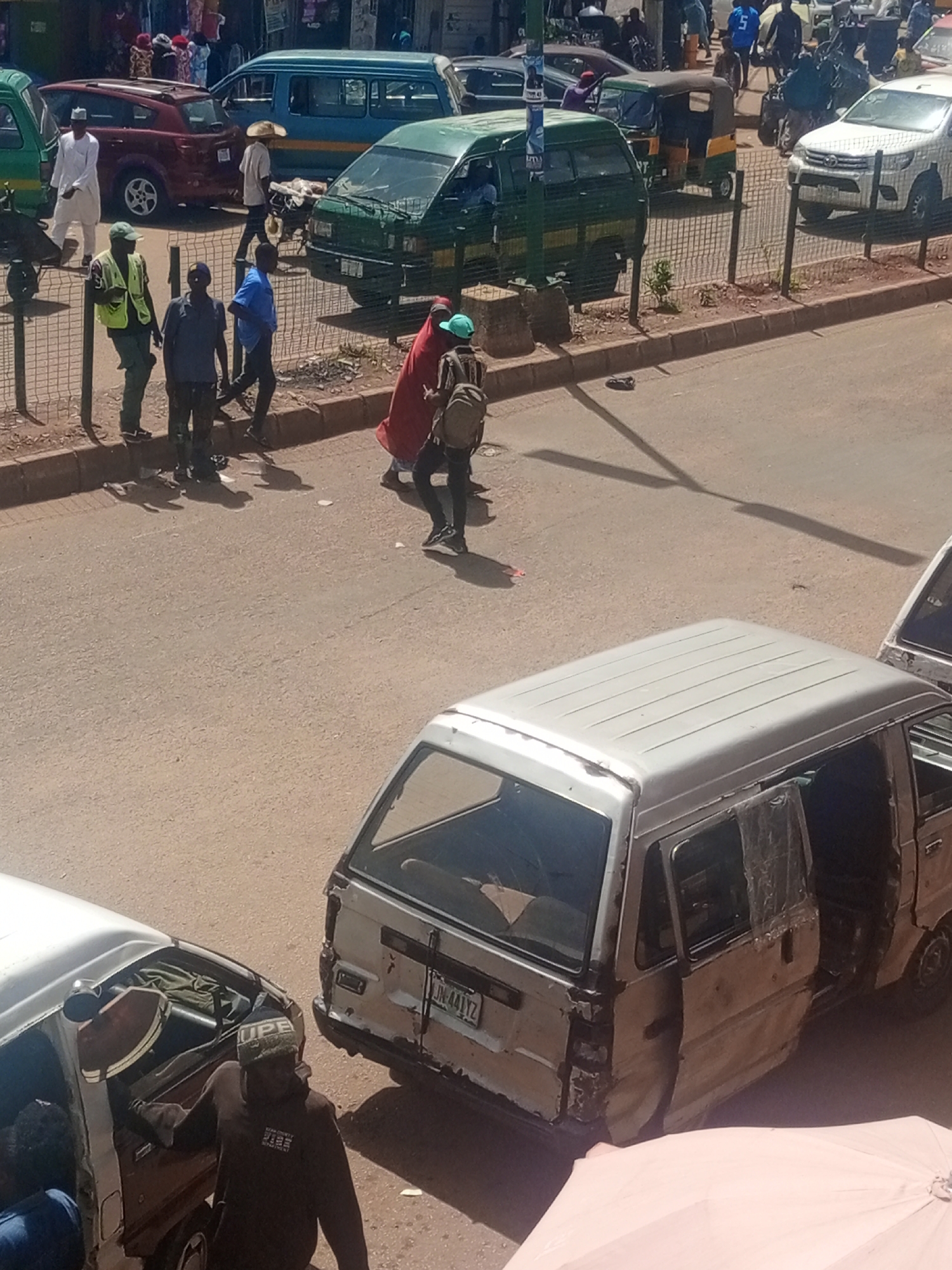In Bukuru, Nigeria, a street scene unfolds with multiple vehicles and pedestrians. A silver minivan, appearing damaged and weathered with open doors and exposed interior components, is parked in the foreground. To its right, another white vehicle, also appearing worn, displays a license plate ending in "441YZ". The street is paved and appears dusty. A group of individuals are standing or walking near a metal fence bordering the road. One person, wearing a blue shirt and dark pants, is walking away from the camera. Another, in a red garment and green headscarf, is walking with a backpack. Several other people are visible in the background, some standing by a maroon car and a blue van. A green commercial van is parked further down the street. The lighting suggests it is daytime, with distinct shadows cast on the road.