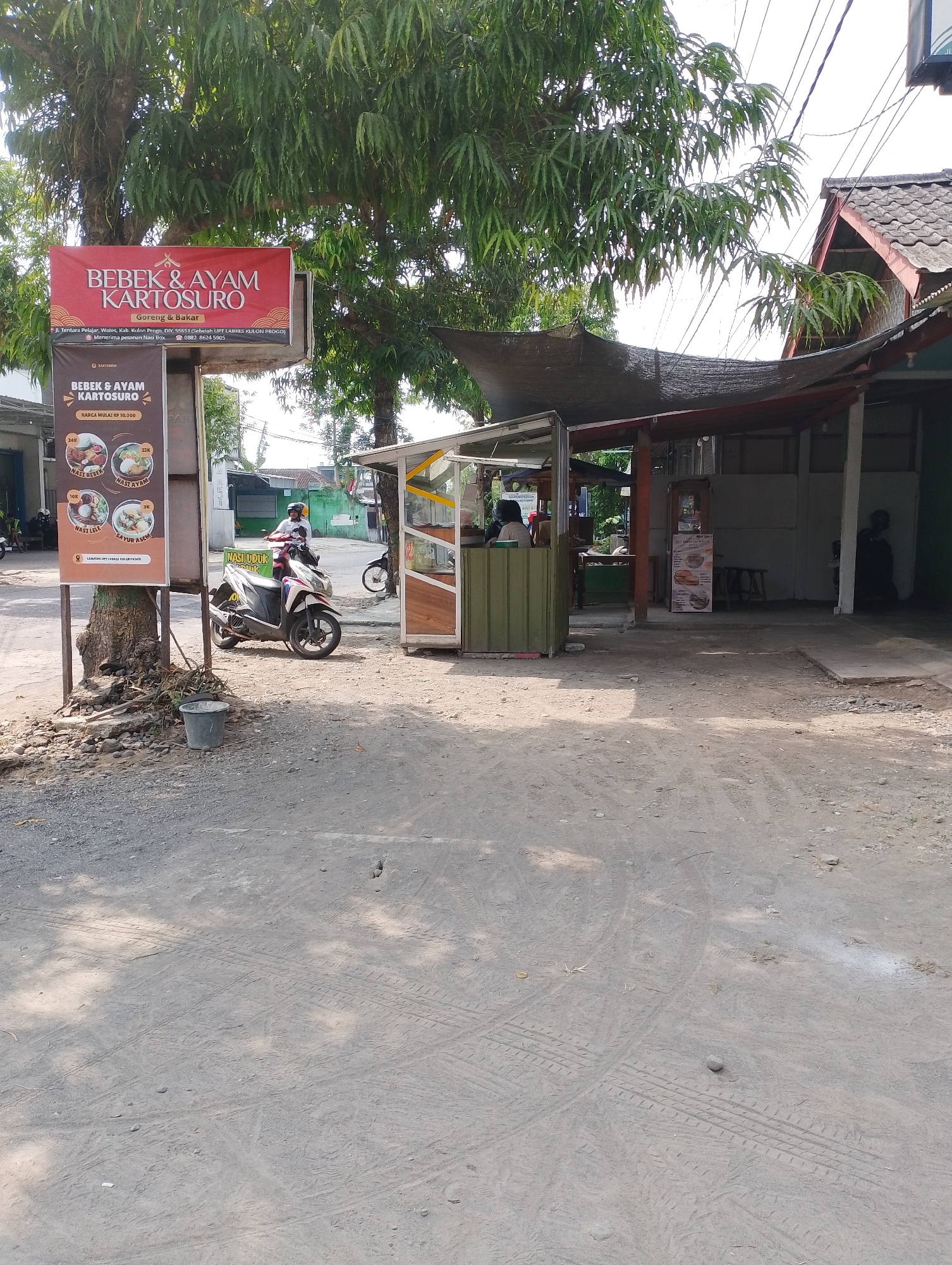 The image depicts a street scene in Wates, Indonesia, likely in front of a food establishment. A prominent red sign with white text reads "BEBEK & AYAM KARTOSURO," indicating it is a duck and chicken restaurant. Below this, smaller text details the address as "JL. Tentara Pelajar, Wates, Kab. Kulon Progo, DIY, 55651 (Sebelah UPF Laires Kulon Progo)" and mentions "Menerima pesanan Nasi Box." A motorcycle, ridden by a person wearing a helmet and a white shirt, is parked in front of the establishment. To the right of the motorcycle is a small food stall with a green and white structure. Further in the background, the roof of a building with traditional Indonesian tiles is visible, along with power lines. The ground in the foreground is unpaved and dusty, with tire tracks visible. Another sign board is located to the left of the red sign, featuring images of food items like "NASI BEBEK" and "NASI AYAM" with prices. The scene is illuminated by daylight, casting shadows.