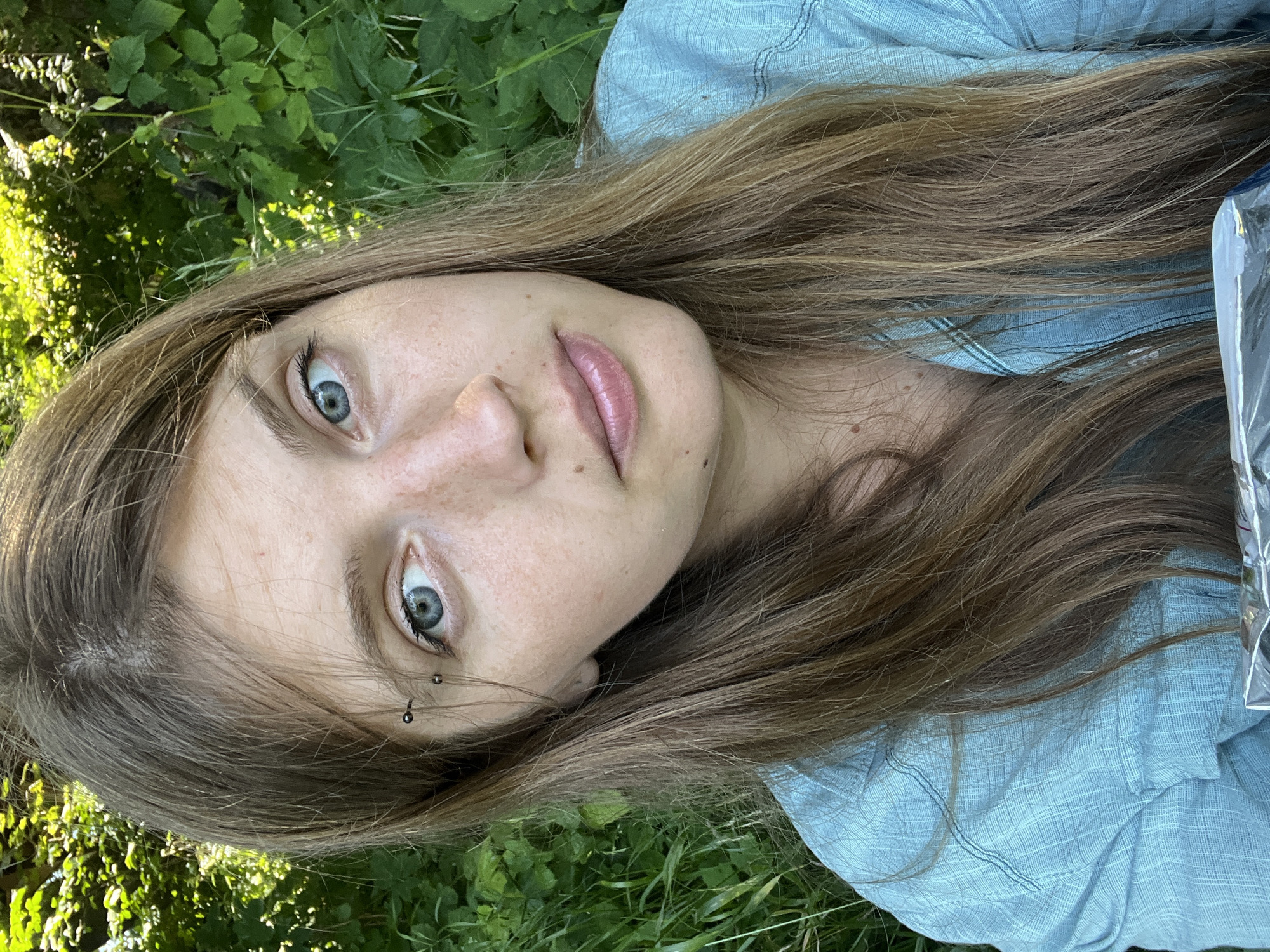 A medium shot selfie captures a young Caucasian woman with long, light brown hair and blue eyes, looking directly at the camera. She has a small eyebrow piercing on her left brow and freckles scattered across her nose and cheeks. She is wearing a light blue, textured button-up shirt. The background is filled with lush green foliage, suggesting an outdoor, natural setting, possibly a park or wooded area, in or around Германавіцкі сельскі Савет, Belarus. The lighting is bright, indicating it is daytime. In the lower portion of the frame, a shiny, reflective object, possibly packaging, is partially visible.