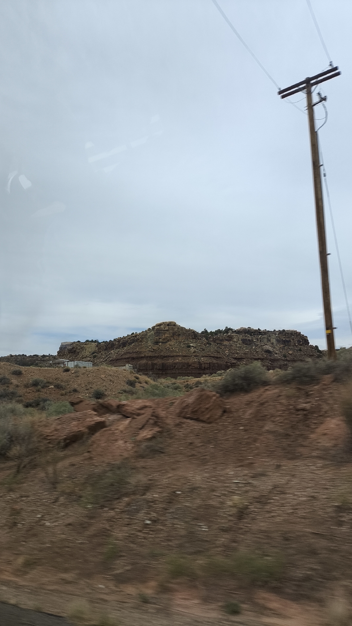 A wooden utility pole stands to the right of the image, supporting two wires that extend across the overcast sky. In the middle ground, a cluster of rock formations forms a layered plateau, with some vegetation growing on top and along its base. At the foot of the plateau, two white buildings with pitched roofs are visible. The foreground consists of a dry, rocky landscape with scattered shrubs and a dark road or surface at the very bottom. The scene appears to be in a rural or semi-arid environment, possibly in Rockville, United States of America, given the geological formations and vegetation. The overall impression is one of a sparsely populated, rugged area under a gray sky.