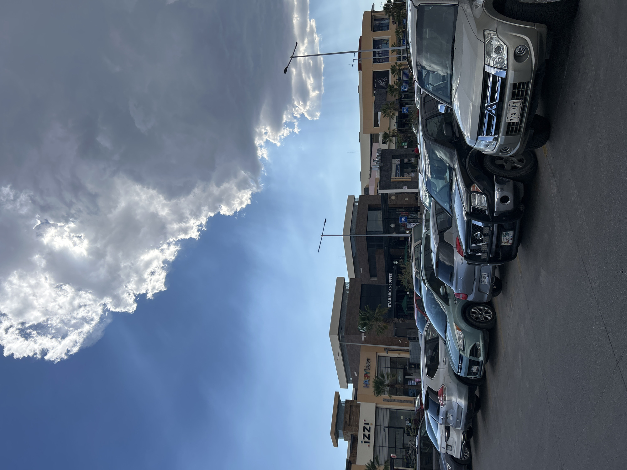 The image shows a street view of a commercial area in Santiago de Querétaro, Mexico. A row of parked cars is visible on the right side of the frame, with the fronts of the vehicles facing the viewer. The buildings on the opposite side of the street house various businesses, including a Starbucks, Happy Laser, and Izzi. Street lights are present. The sky is partially cloudy, with a bright blue section visible amidst the clouds. The license plate of the lead car is 774-ZHY.