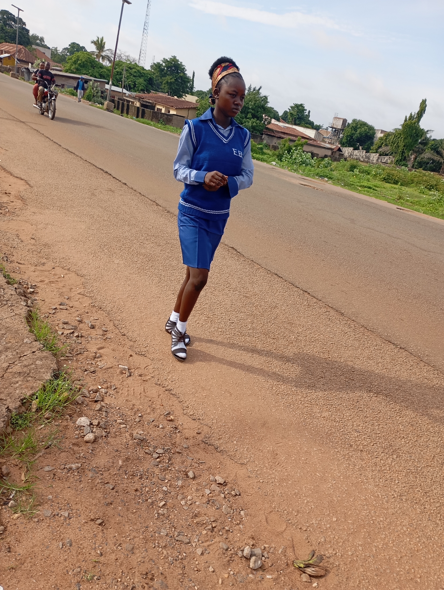 A young woman in a blue school uniform, complete with a blue vest and a blue skirt, is captured mid-stride on an asphalt road in Shendam, Nigeria. The uniform features white trim and the initials "E.B." are visible on the vest. She wears white socks and black and white sandals. Her head is adorned with a multi-colored patterned headband. The road has a distinct beige color, characteristic of many Nigerian roads, and is bordered on the left by a dirt embankment with scattered rocks and sparse green grass. In the background, a motorcycle carrying two individuals is visible in the distance, along with a power line pole, and various buildings and lush green trees that suggest a semi-urban or rural setting. The sky is partly cloudy. The overall lighting indicates it is daytime.