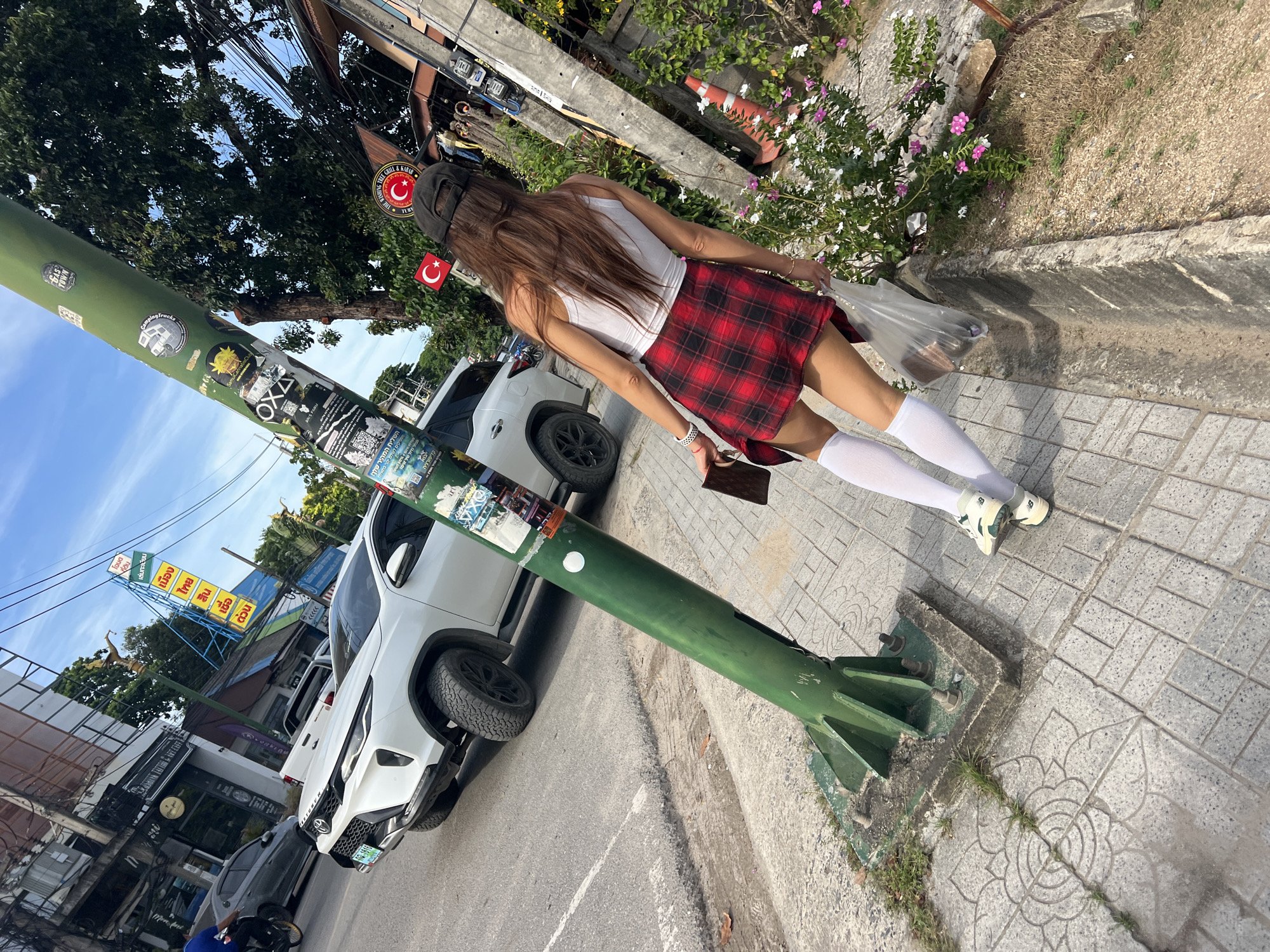 The image is taken in Baan Bang Rak, Thailand. It shows a woman walking away from the viewer on a sidewalk next to a street. She is wearing a white tank top, a red plaid skirt, white knee socks, and light-colored sneakers. She is also wearing a black baseball cap and carrying a clear plastic bag in one hand and a brown object, possibly a wallet, in the other. A green pole with stickers and signs on it stands prominently on the left side of the image. Behind the pole, there's a white SUV parked alongside the road. In the background, there is a building with a vertical yellow and blue sign. Other parked cars are visible in the distance. The sky is partly cloudy. Flowers bloom along the edge of the sidewalk.