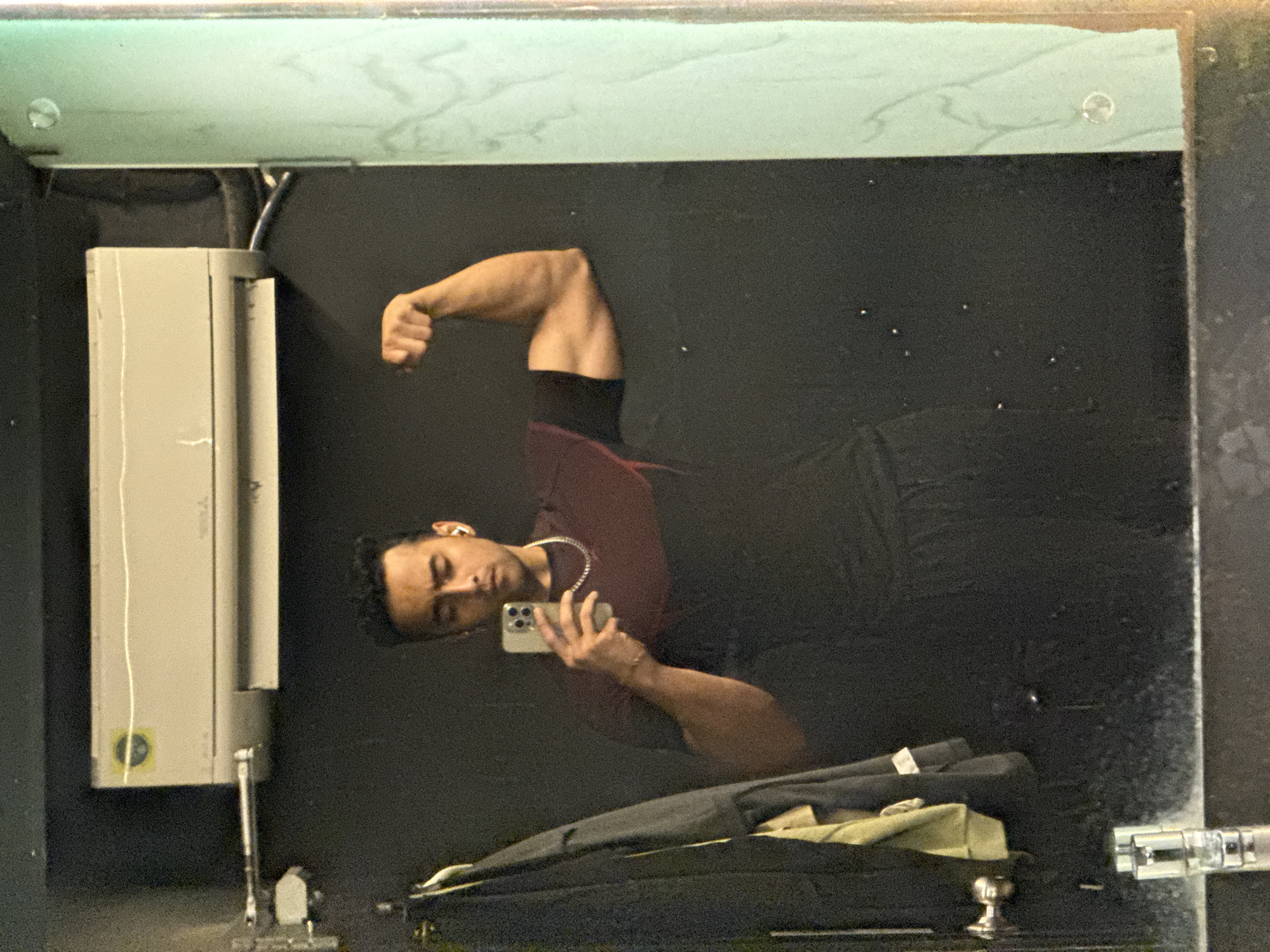A man stands in front of a mirror in a room. He is flexing his left arm and holding a mobile phone in his right hand, presumably taking a picture of himself in the mirror. He wears a dark red t-shirt and a silver chain. A hand dryer is affixed to the wall to the left of the mirror. A dark-colored garment is draped beneath the mirror. The exact location in Kolkata, India, is impossible to determine from the visual information.