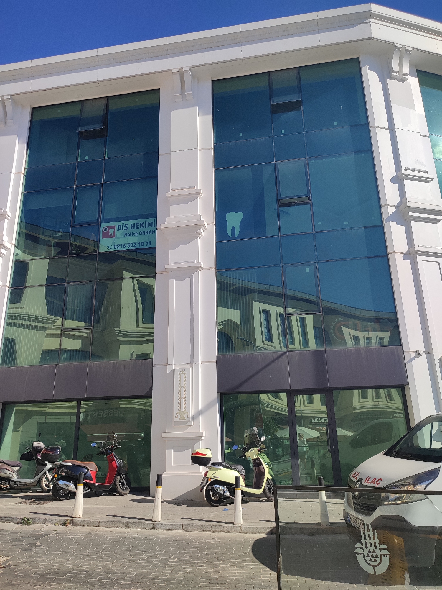 This is a daytime, eye-level exterior shot of a modern, multi-story building with a glass facade, located in Üsküdar, Turkey. The building features large, blue-tinted windows that reflect the surrounding environment. A white sign is visible on the upper floor, indicating a dental practice. Below the glass facade, on the ground floor, are large glass doors and windows. A street runs in front of the building, paved with light-colored cobblestones. Several parked scooters and a white van with the text "ACİL İLAÇ" (Emergency Medicine) are present on the sidewalk in front of the building. The sky is clear and blue.
