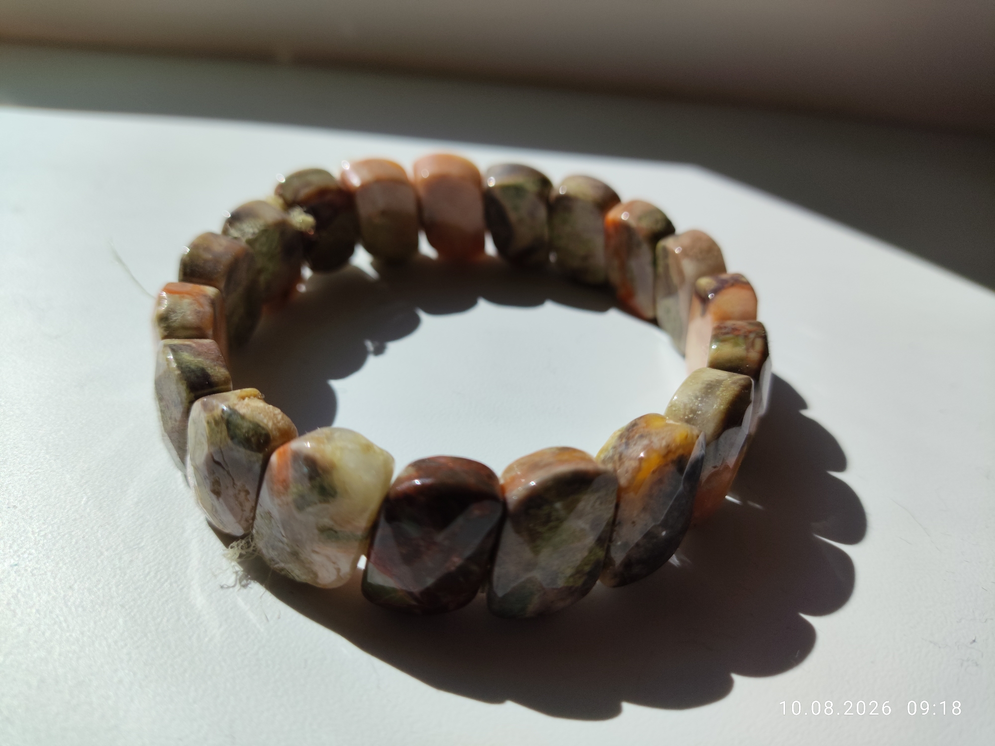 An unbranded, faceted stone bead bracelet is displayed on a white surface. The bracelet is circular in shape, with the beads showing a mix of earthy tones including browns, greens, and peachy oranges. The beads are irregular in shape but polished to a shine, with each bead cut with multiple facets that catch the light. The bracelet is illuminated by sunlight from the left, casting a shadow with scalloped edges to the right. A date and time stamp, "10.08.2026 09:18", is visible in the lower right corner of the image. The overall environment appears to be indoors, near a window, with the white surface possibly a table or windowsill. There are no people or any other objects present in the scene. The location is not specified.