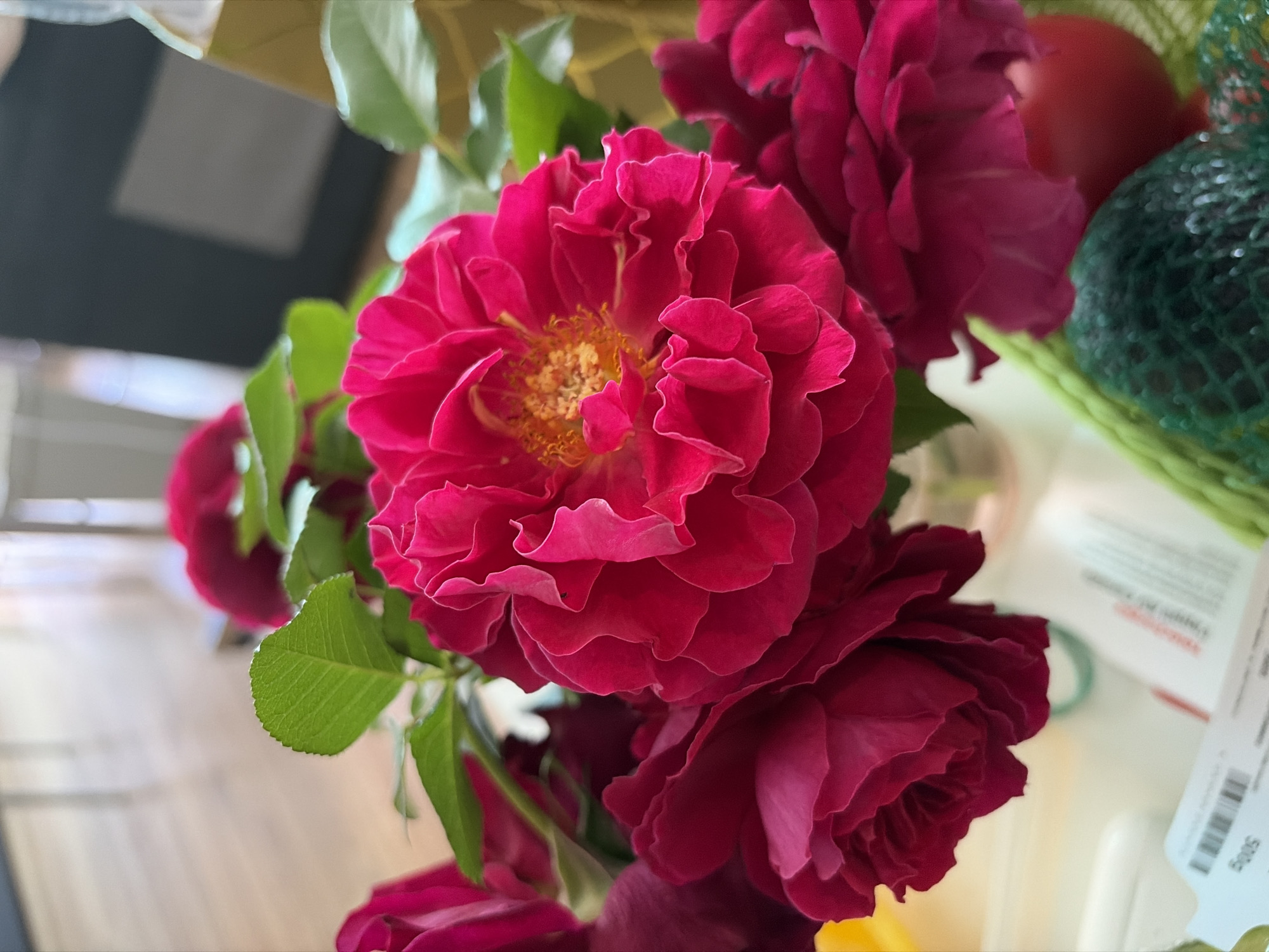 A close-up shot focuses on a bouquet of vibrant pink roses, with a prominent, fully bloomed rose in the center. The petals of the central rose are intricately layered and ruffled, revealing a cluster of yellow stamens at its core. To the left, a darker pink rose is partially visible, its petals also showing a rich, velvety texture. The bouquet is adorned with lush green leaves. In the background, the scene is softly blurred, suggesting an indoor setting with natural light illuminating wooden flooring. A dark object with a lighter square on it is visible, along with a portion of a glass bottle containing a yellowish liquid. At the bottom of the image, a green mesh bag and a red sphere are partially visible, along with a white object that appears to be packaging with some text. The overall impression is one of a domestic or artistic setting, potentially a home or a studio in Lublin, Poland.