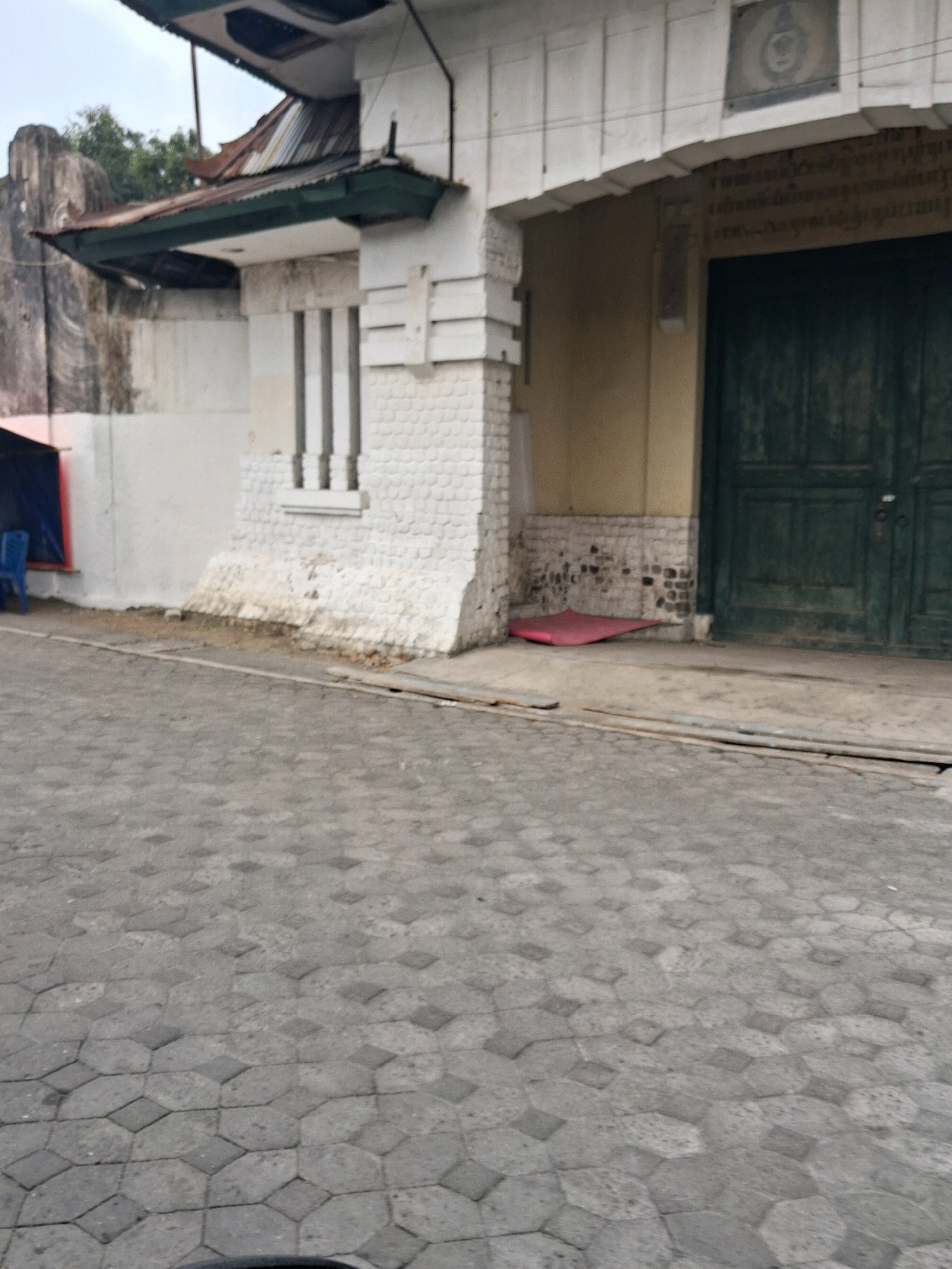 A view of a weathered building with a large green door and a cobbled street in front of it. The building has a white facade with decorative elements and a corrugated metal roof. There is a blue chair and a red mat visible near the entrance. The street is paved with hexagonal gray stones. The image appears to be taken in the city of Surakarta, Indonesia, likely in an older district given the architectural style and materials.