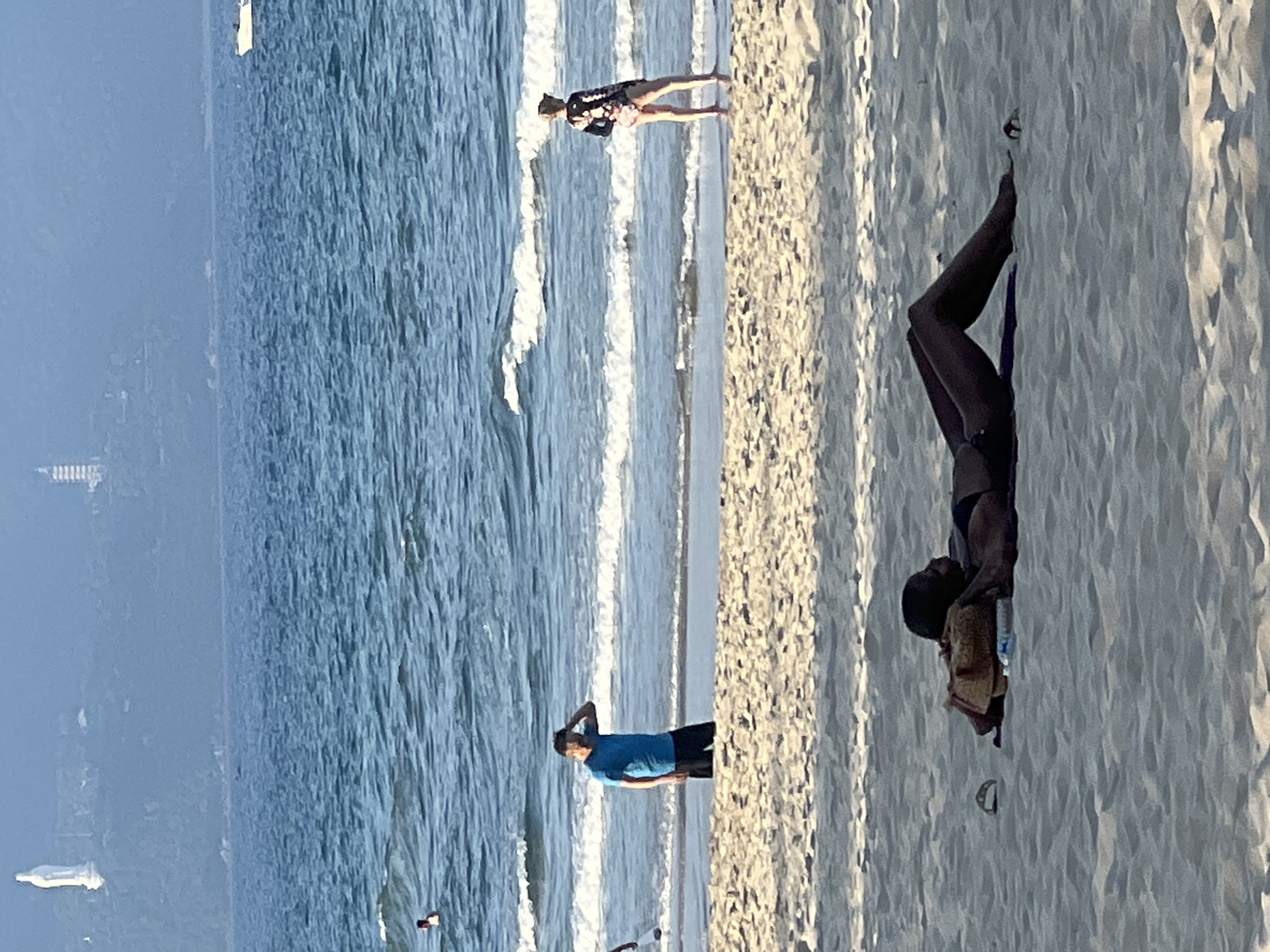 The image depicts a beach scene in Đà Nẵng, Vietnam. In the foreground, a person is lying on the sand, seemingly relaxed. In the mid-ground, two individuals are present on the beach near the water's edge. One person, wearing a blue t-shirt and dark shorts, is standing and appears to be shielding their eyes. The other person is wearing a patterned swimsuit and appears to be looking out at the water. The sea stretches out in the background, with gentle waves lapping the shore. Far in the distance, on the hills overlooking the city, a large white statue of a woman is visible, along with other structures. A boat can be seen in the far distance on the water. The overall impression is one of a tranquil day at the beach.