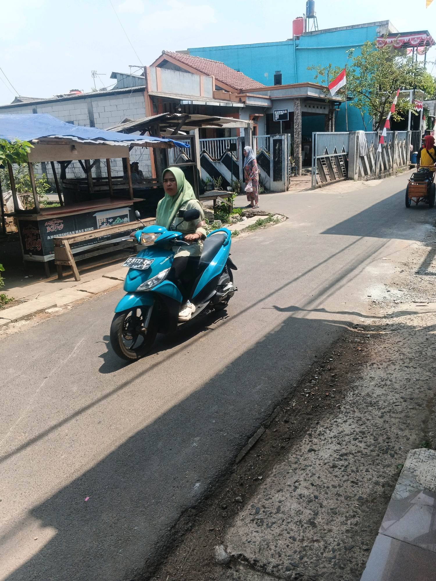 A woman wearing a light green hijab and a light green jacket is riding a bright turquoise scooter down a paved street. The scooter has a license plate that reads "G 8172 BW". She is in motion, and her shadow is cast across the road. To the left of the street, there is a covered market stall with a blue tarp roof and wooden supports. The stall has shelves and appears to be empty. Further in the background, there are buildings with red-tiled roofs and a bright blue building with a red water tank on top. Indonesian flags are visible on some of the buildings. To the right of the street, there is a fence and another person is partially visible with a cart. The scene appears to be in a residential or commercial area in Kadu, Indonesia, under a bright, sunny sky.