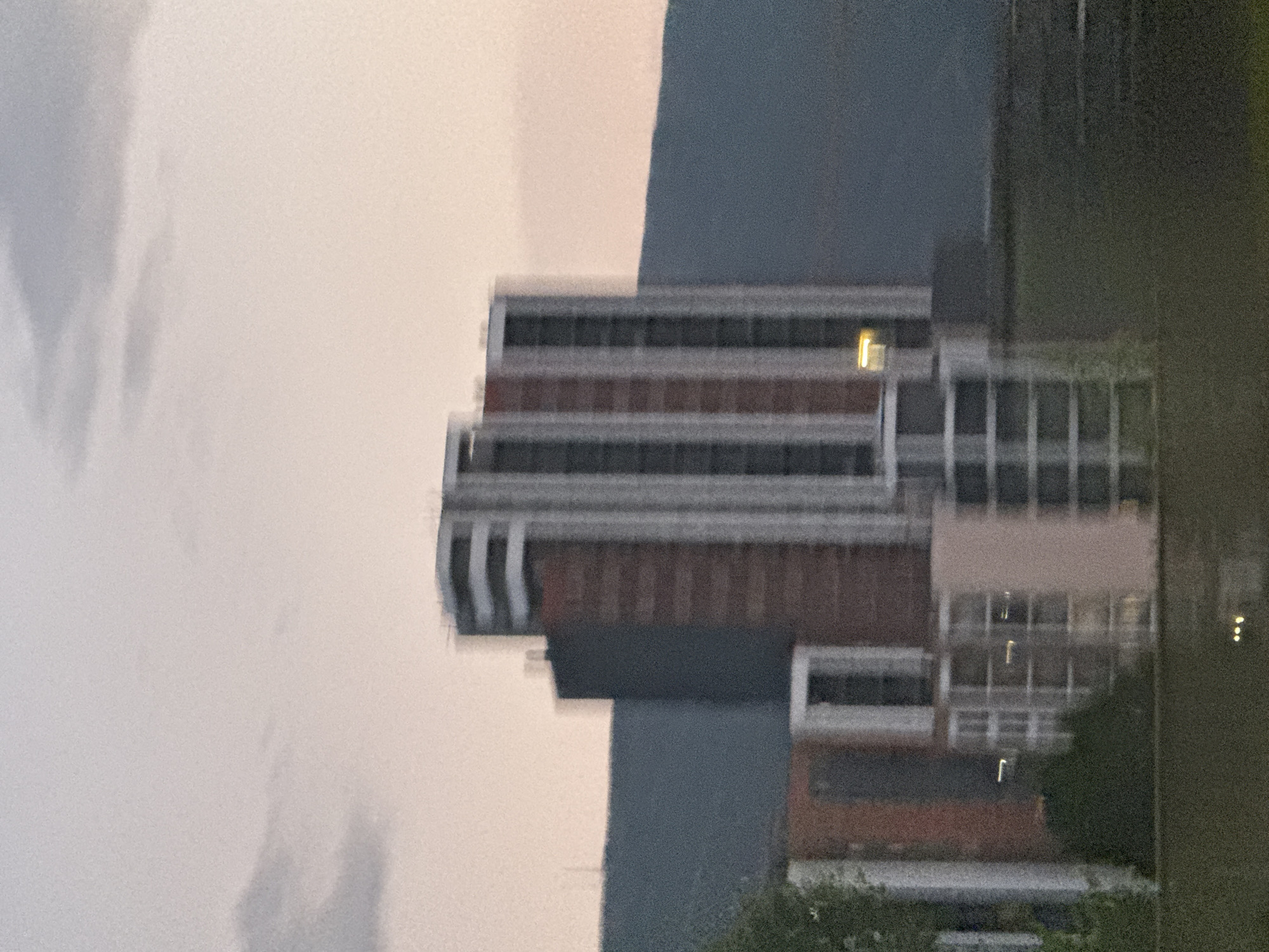 The image depicts a collection of modern buildings in Tbilisi, Georgia, set against a hazy, pale sky and a dark, blurred mountain range. The buildings appear to be residential or office complexes, characterized by varying heights and architectural styles. One prominent structure features a reddish-brown brick facade with tall, vertical white columns or balconies. Adjacent to it is a taller building with a grey and white striped exterior, featuring multiple floors with visible windows. To the left and in the foreground, other buildings with lighter facades and distinct window patterns are visible, some with orange accents. The overall scene is softly lit, suggesting it was captured during either dawn or dusk, contributing to the blurred and muted tones of the image. The foreground shows a dark, indistinct area that could be water or a grassy expanse, with small, faint lights reflected on its surface.