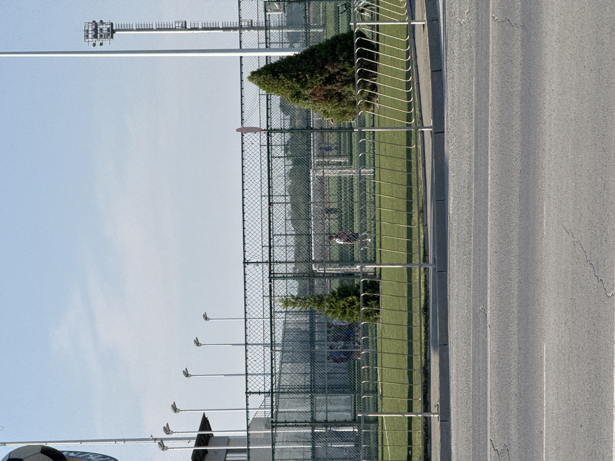 This is a photograph of a sports field in Yevpatoriya, Ukraine, on a clear, sunny day. The field is enclosed by a chain-link fence. In the foreground, a paved road runs horizontally across the bottom of the image, separated from the field by a curb and a metal barricade. Beyond the fence, the green turf of a soccer field is visible. Several streetlights stand tall around the perimeter of the field.
On the field, a group of young people are engaged in what appears to be a soccer practice. They are dressed in athletic clothing and are scattered across the field, some with a ball. One person is positioned near a goal. A small evergreen tree stands to the right of the frame, casting a shadow on the field. In the background, behind the fence, are buildings and more trees, suggesting an urban or suburban setting. A tall utility tower with lights is visible in the far right of the image.