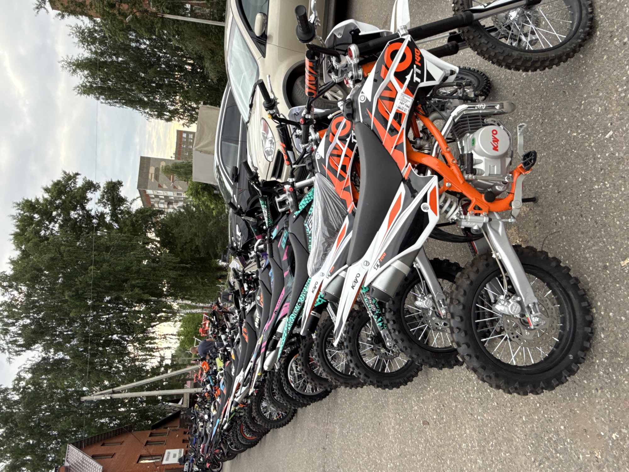 A fleet of dirt bikes are parked in a row on a paved surface, likely a street or parking lot, in Votkinsk, Russia. The bikes are predominantly orange and white, with black accents and some featuring colorful graphics. Several bikes have their wheels turned to the left, suggesting they are parked facing forward. The bikes are lined up closely, with their front wheels almost touching the rear of the bike in front. In the background, a brick building with a pitched roof is visible on the left, and several trees with dense green foliage stand tall. Beyond the trees, urban buildings, including a multi-story apartment building, are discernible, under a cloudy sky. The foreground is dominated by the tires and lower chassis of the closest dirt bikes. A single person, a male with short hair and wearing a dark t-shirt, is visible standing among the bikes in the middle ground, facing away from the camera. The overall scene depicts a collection of off-road motorcycles prepared for display or sale in an outdoor urban environment.