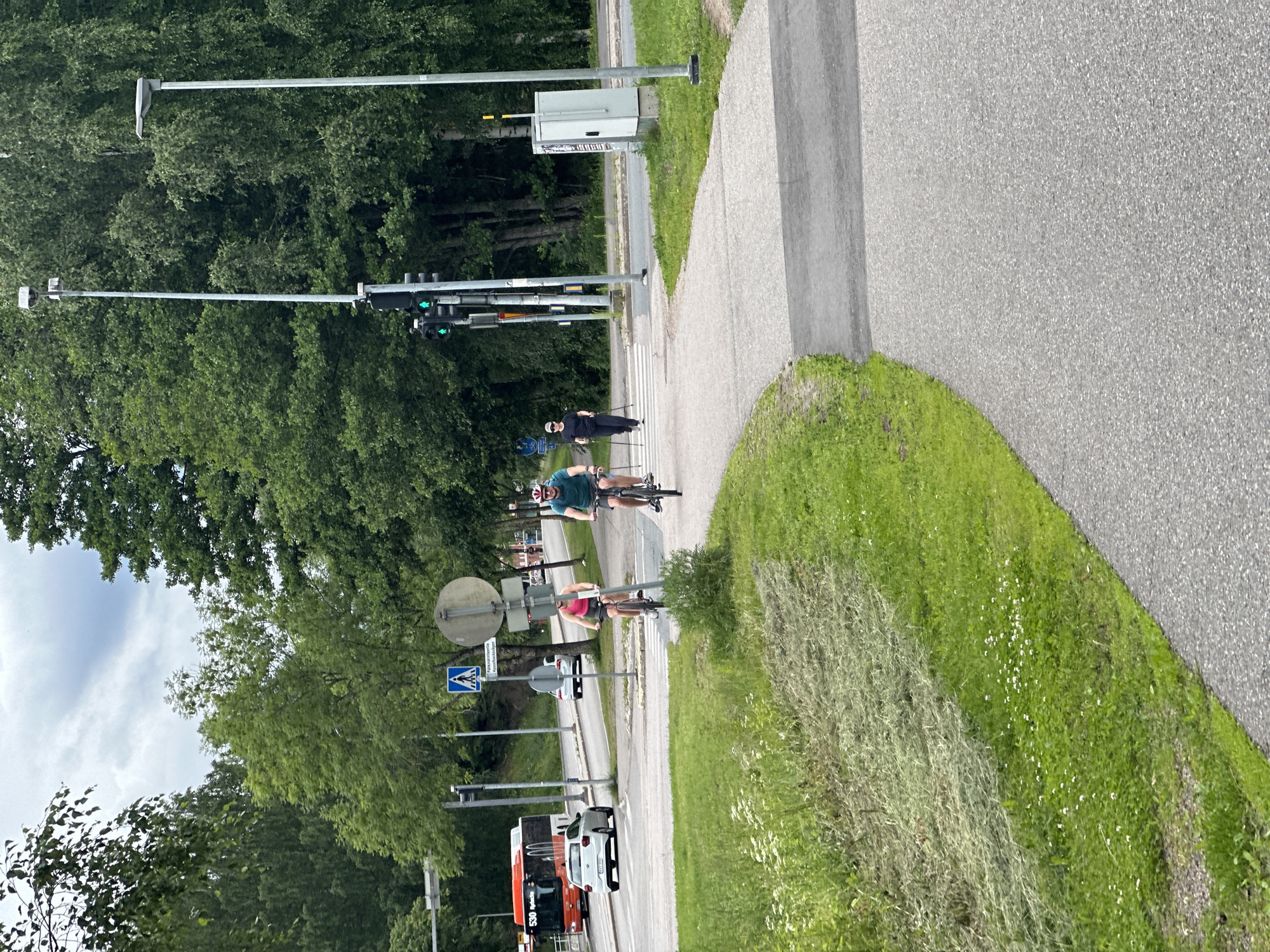 The image depicts a street scene with people, vehicles, and traffic signals. The primary subjects are two individuals cycling on a paved path adjacent to a road. One cyclist is wearing a blue t-shirt and black shorts, and the other is wearing a red top and dark shorts. Both are on bicycles.
The scene is set outdoors, with lush green trees lining the left side of the road. A paved path runs parallel to the road, separated by a grassy verge. On the right side of the path, there's a sloping grassy area with some wildflowers and dry grass. The road itself is grey asphalt.
In the background, several traffic lights are visible, some showing green signals. There's also a white pedestrian crossing marked on the road. Further down the road, a white car and an orange bus are parked. The sky appears overcast with some white clouds.
Notable details include the presence of traffic infrastructure like poles and signal boxes. The overall impression is that of a pleasant daytime scene, possibly in a suburban or park-like setting, given the abundant greenery. There is no clear indication of the time of day or the specific location. No visible emotions are discernible from the distant figures.
There are some signs visible, including a blue square sign with a white pedestrian symbol, indicating a pedestrian crossing. Another circular sign with some text is also present but unreadable. The bus has "530" and some text in a foreign language, possibly "Päivänniemi," visible on its side.
