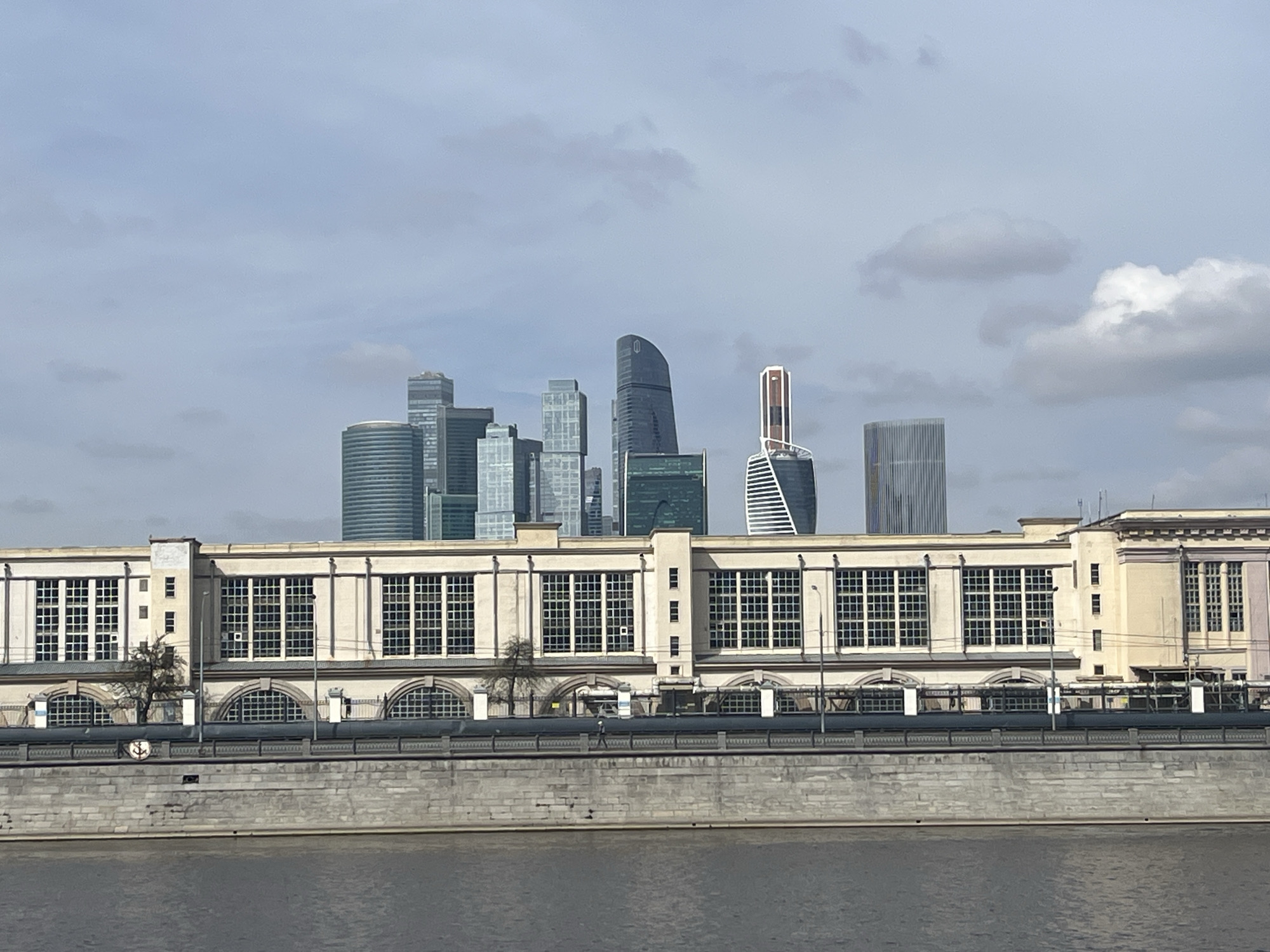 The image shows a cityscape scene in Moscow, Russia, featuring a large, beige building in the foreground and several modern skyscrapers in the background. The building has a symmetrical design with numerous windows, arches, and a flat roof. In front of the building is a gray concrete embankment, followed by a body of water, likely the Moskva River. The skyscrapers in the background vary in height and design, some with rounded tops and others with sharp edges. The sky is overcast with gray and white clouds. There is a person walking in front of the building.