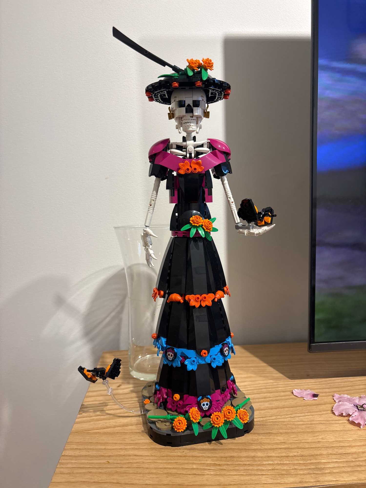 A Lego build of a skeletal figure in a traditional Mexican Day of the Dead costume, known as a Catrina, stands upright on a wooden surface. The figure is adorned with a wide-brimmed black hat decorated with orange flowers and green leaves, a pink ruffled collar, and a long, black, tiered dress featuring colorful floral and skull motifs. The figure's arms are articulated, with one hand holding a small black and orange Lego creature. Two small Lego butterflies are positioned on the base of the display, one attached to a clear stand. In the background, a white wall casts a shadow, and to the right, a television screen displays a colorful image. A clear glass vase is partially visible behind the figure. Scattered on the wooden surface to the right of the figure are two light pink flower petals. The scene is indoors and appears to be a domestic setting, possibly a shelf or tabletop. There is no visible indication of the location being in Buenos Aires, Argentina.