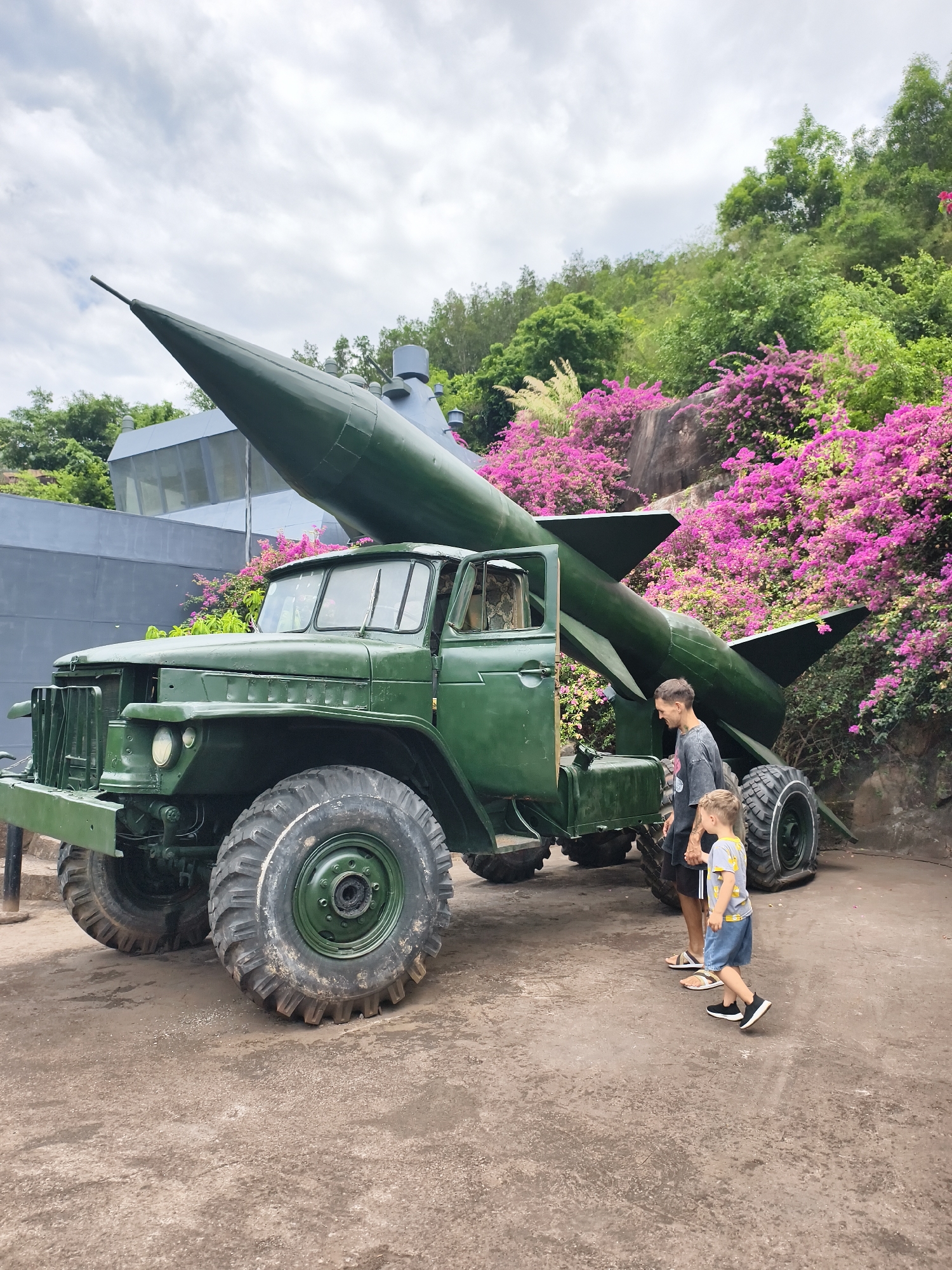The image shows a green missile launcher truck parked on a paved surface in what appears to be a park setting. A man and a young child are standing near the truck. The truck has large, heavy-duty tires, and a green missile is mounted on the back. Pink bougainvillea flowers are visible in the background, along with green trees and a building. The image was taken in Tay Nha Trang Ward, Vietnam.