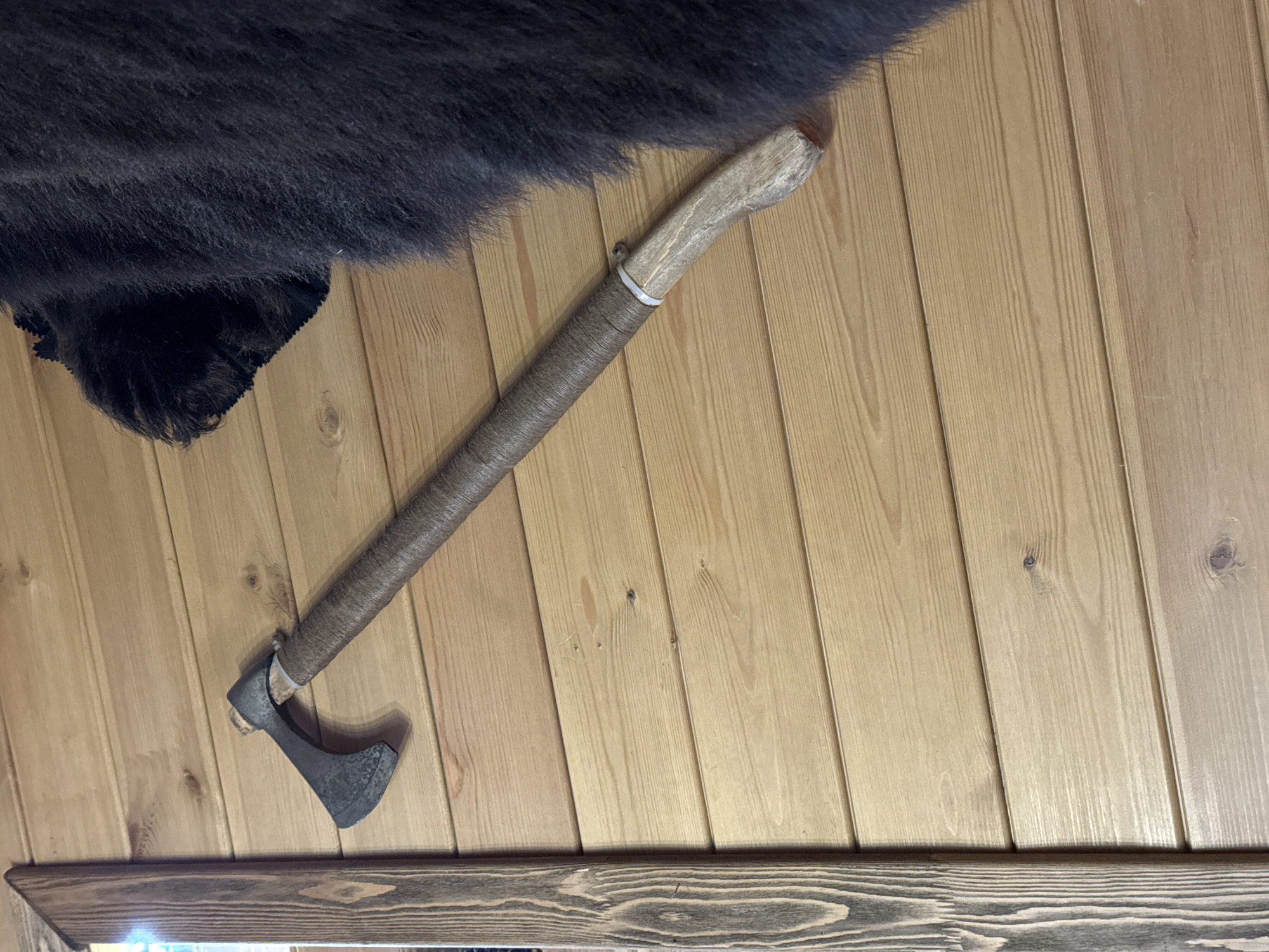 A wall made of vertical wood planks is adorned with a bear pelt and an axe. The handle of the axe is wrapped with a tan material. A dark, wooden shelf is located near the bottom of the image. The location is Ново-Никольское, Russia.