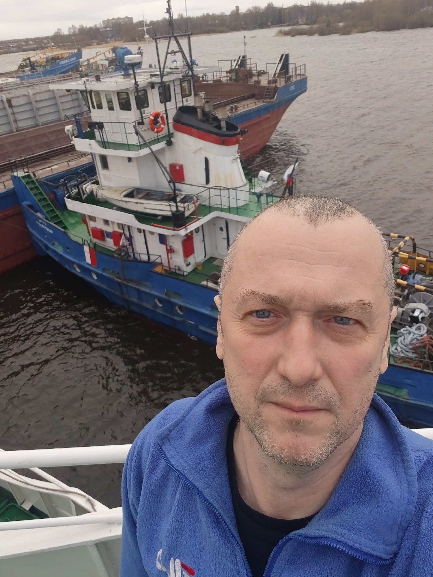A man takes a selfie on what appears to be a ship's deck, with other vessels and a river scene in the background on an overcast day in Bol'shie Mkhi, Russia. The man, in his late 40s or 50s, has a receding hairline, blue eyes, and light stubble. He wears a blue fleece jacket with a partially visible logo on the left chest, possibly "UHF," over a dark shirt. His expression is neutral.
Behind him and to his right, a blue and white tugboat is prominently featured on the water. Its superstructure is white with green trim, and a red lifebuoy hangs on its side. A small white dinghy is stowed on its deck. Visible on the hull of this boat is the text "ОТТОН СВОРОВСКИ" (OTTON SVOROVSKI).
Further back and to the left of the tugboat, a larger, red and blue barge or cargo vessel is partially visible, likely being pushed or towed by the tug. The text "ТИТАН" (TITAN) is legible on its bow. In the far background, across the water, another blue vessel can be seen, with the partially visible text "СОЮЗ..." (SOYUZ...). The river surface shows ripples, indicating movement or current. The distant shoreline features trees and some indistinct buildings under a cloudy sky. The man is likely on a larger vessel, looking down at the working boats below.