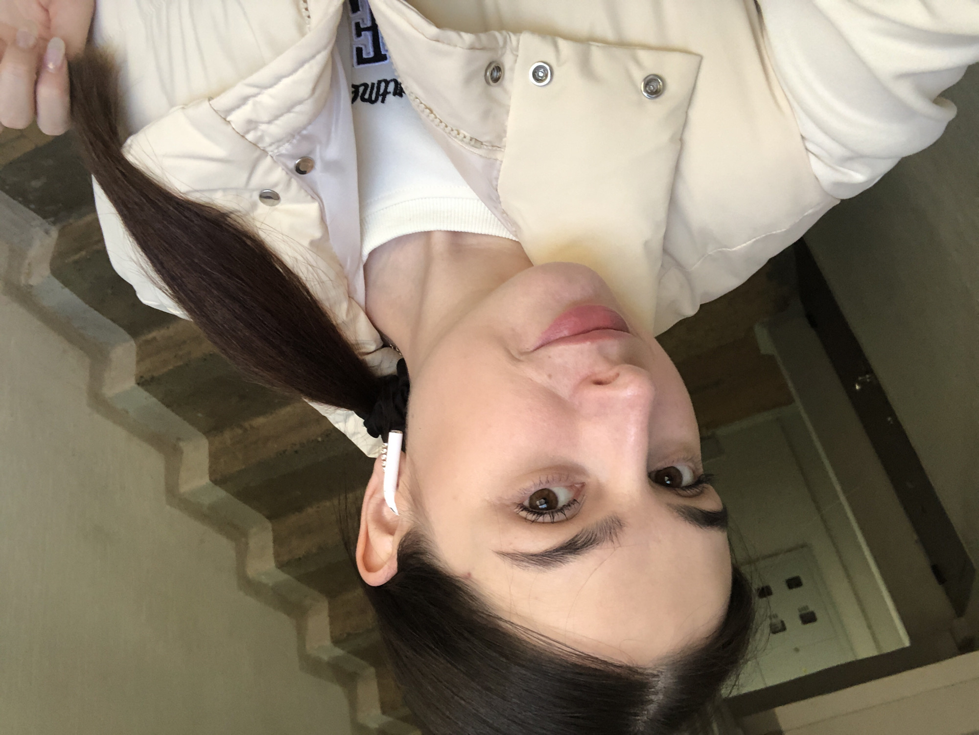 A woman with dark hair pulled into a ponytail stands indoors. She wears a cream-colored puffer vest over a white top. She has an earbud in her ear. Behind her are stairs. A light switch is visible on the wall. The image was taken upside down. There is insufficient information to confirm the location is Chaikovsky, Russia.