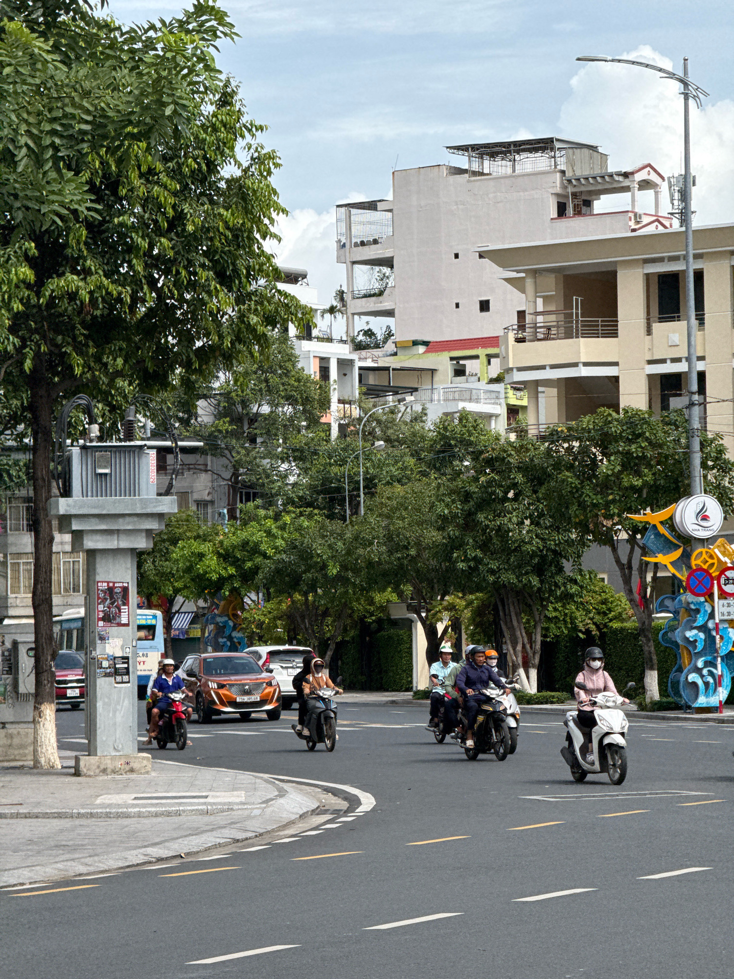 This is a street scene in Nha Trang, Vietnam, with a cloudy sky. The road is paved with asphalt and has white and yellow lane markings. Several motorbikes are on the road, some with riders wearing helmets and masks. There are also cars and a bus visible. Buildings line the street, some with multiple stories and balconies. Trees with green foliage are scattered throughout the scene, providing shade. A street light and utility pole are also present. A decorative sculpture with a circular sign is visible on the right side of the image.