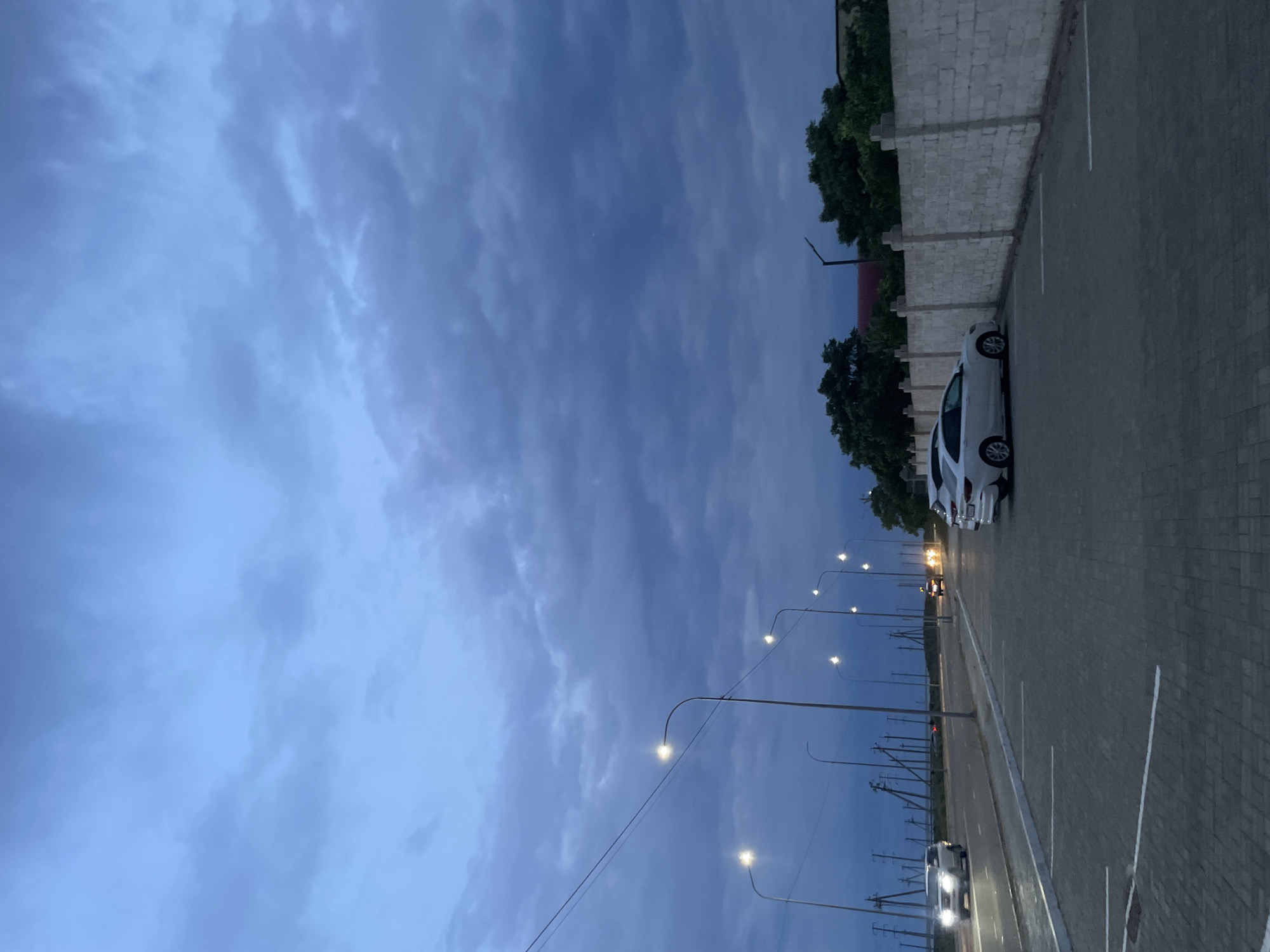 The image depicts an outdoor scene at dusk in Aktau, Kazakhstan. The sky is filled with dark, heavy clouds, indicating an overcast or approaching storm. A row of streetlights illuminates a paved parking lot in the foreground, with white parking lines visible. Several white cars are parked in the lot, and one car is driving on an adjacent road, with its headlights on. In the background, a low, light-colored brick wall runs along the edge of the property, topped with cylindrical posts. Beyond the wall, dense green trees and a few buildings are visible. The overall atmosphere is somber and moody, characteristic of twilight in a coastal city.