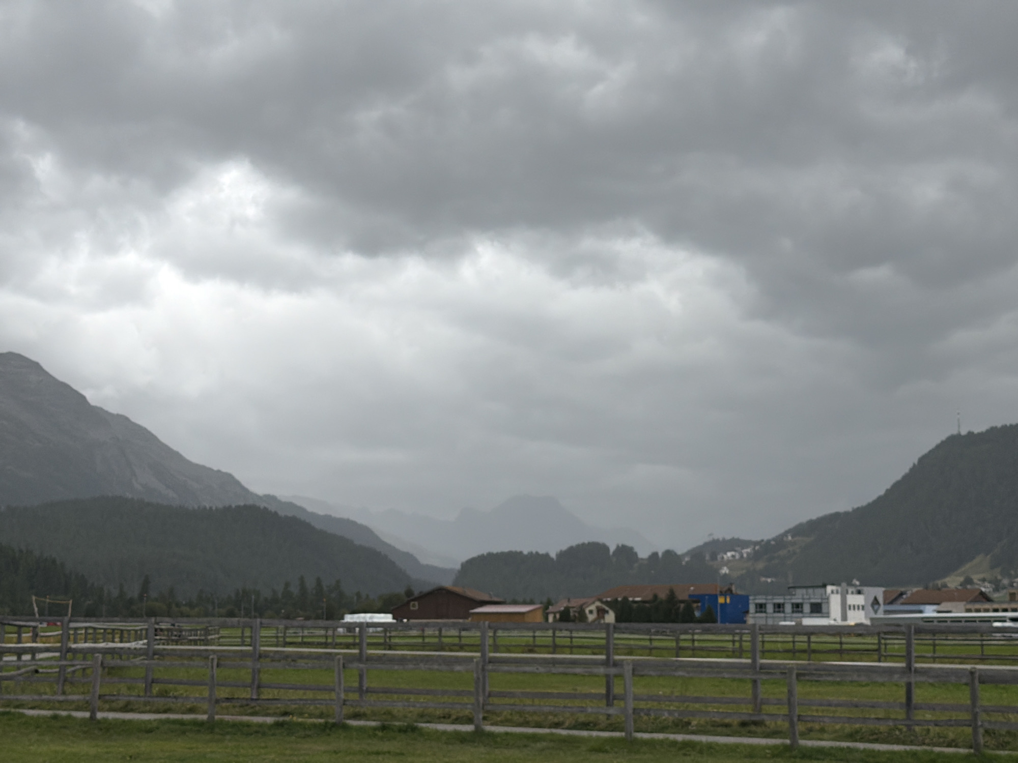 The image depicts a wide shot of a rural landscape under a heavily overcast sky, possibly indicating impending rain or a recent downpour. In the foreground, a grassy field is bordered by a wooden fence constructed from horizontal planks and vertical posts. Beyond the fence, a track or path runs parallel to the fencing.
Further into the mid-ground, a series of agricultural or equestrian facilities are visible. These include several low-rise buildings with reddish-brown or white facades, some with visible roofs. One building appears to be a stable or barn, while others suggest commercial or residential use. There are also fenced enclosures, likely paddocks or fields for animals. A tall, yellow net structure, possibly for a sport like volleyball or tennis, is present on the left side of the mid-ground.
The background is dominated by a series of rolling, tree-covered hills that recede into the distance, culminating in rugged, mist-shrouded mountains. The overall atmosphere is subdued and possibly damp, with the gray clouds casting a diffused light over the scene. There are no people or animals visible in the image. The location is identified as Samedan, Switzerland.