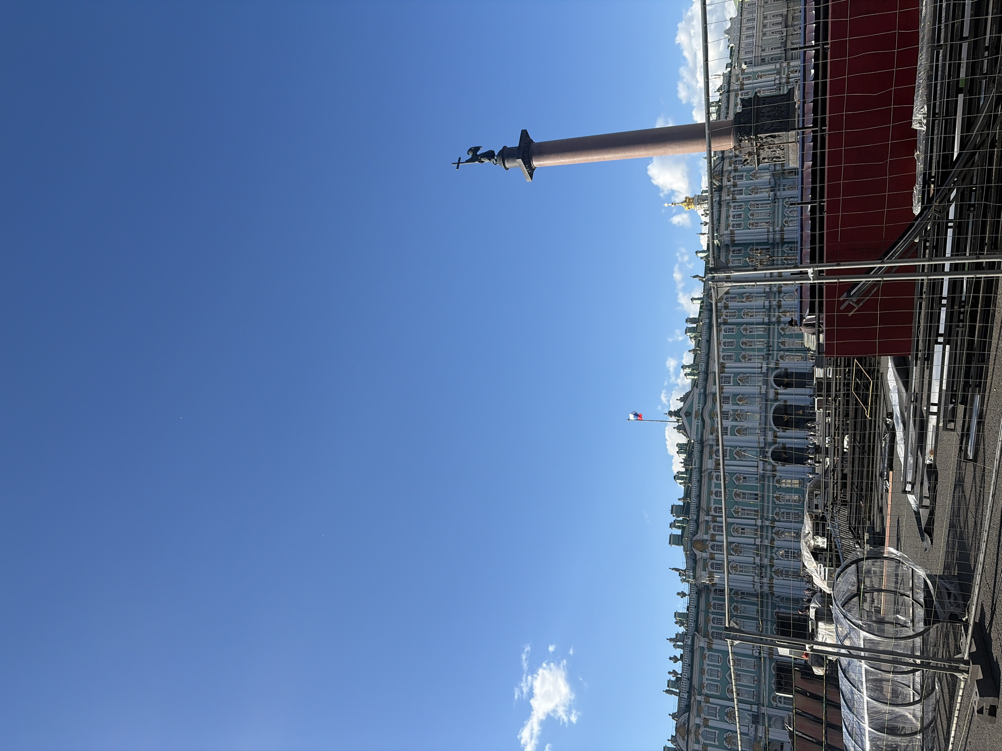 The image captures an outdoor scene in Palace Square, Saint Petersburg, Russia, dominated by the expansive blue sky and a section of the grand Winter Palace, painted in a distinctive pale turquoise with white neoclassical accents. In the foreground, a temporary construction fence encloses an area with various materials, including metal scaffolding, plastic sheeting, and a cylindrical transparent structure. The fence itself is made of metal mesh panels.
To the right of the Winter Palace, the iconic Alexander Column stands tall against the sky. It's a tall, slender, reddish-brown granite column topped with a statue of an angel holding a cross. A small Russian flag is visible atop a building to the left of the column, fluttering in the breeze.
People are sparsely visible in the background, some walking in front of the Winter Palace. The overall impression is one of a public space, possibly undergoing some form of preparation or renovation, with the historical grandeur of the architecture providing a striking contrast to the modern fencing and construction elements. The lighting suggests it is daytime, with clear skies and a few scattered white clouds.