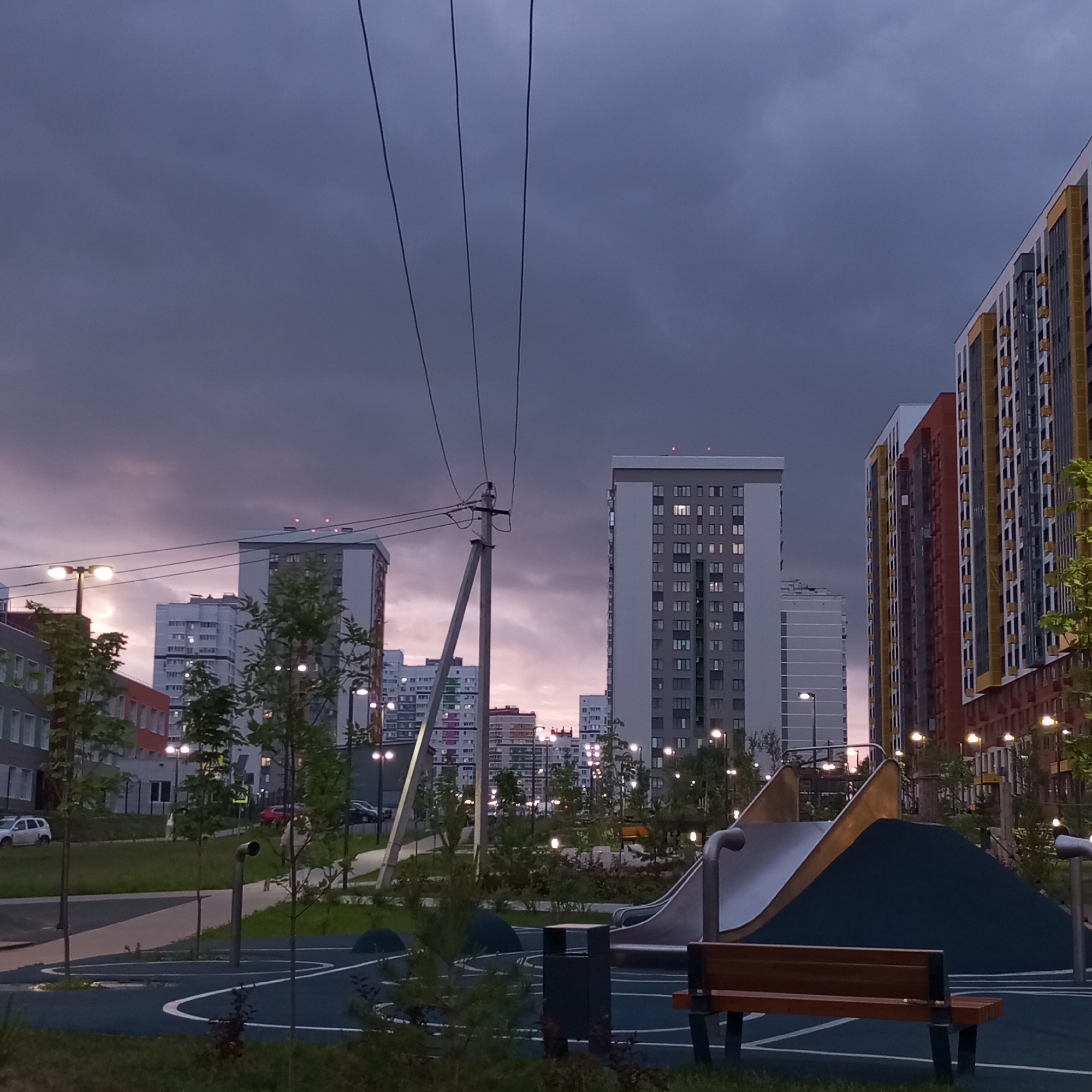 The image depicts a modern residential area in Izhevsk, Russia, during twilight. In the foreground, a playground features a slide, a wooden bench, and a trash receptacle, all situated on a blue rubberized surface with white markings. Several young trees are scattered across the scene. In the mid-ground, lampposts illuminate the area, casting light on pathways and surrounding greenery. Various multi-story apartment buildings are visible in the background, with lights on in some windows. Overhead, thick power lines stretch across the frame against a dark, cloudy sky tinged with the colors of dusk. Several cars are parked on the street in the distance.