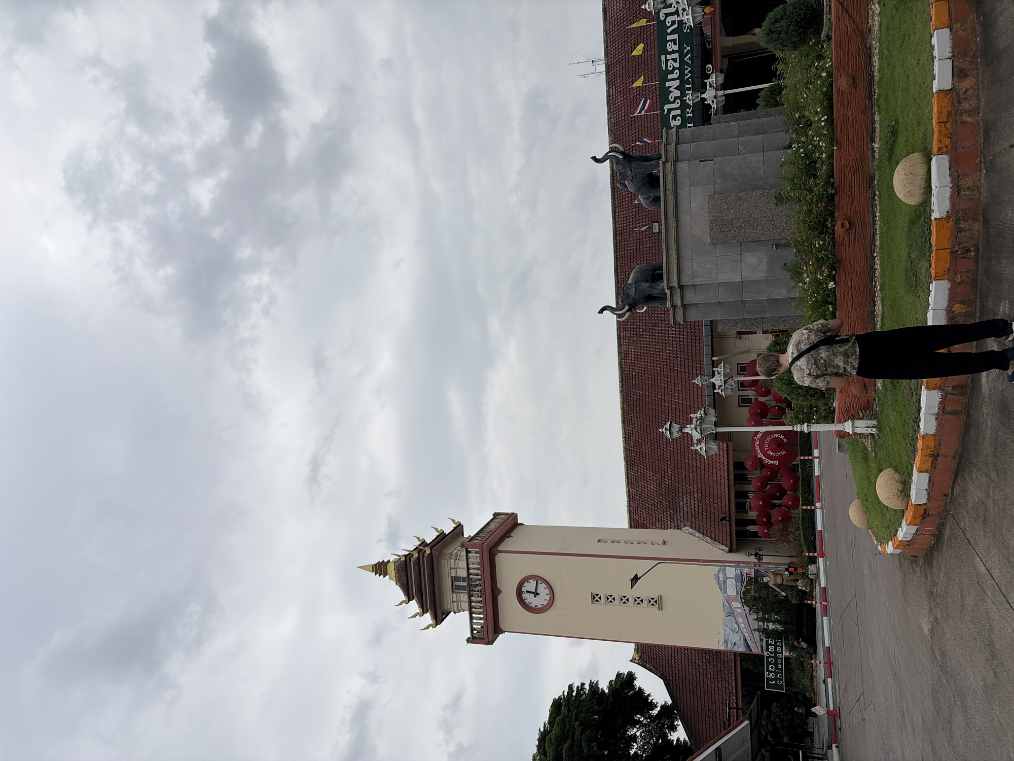A medium shot captures a view of the Chiang Mai Railway Station in Chiang Mai City Municipality, Thailand, on an overcast day. The station's architecture features a prominent clock tower with a tiered, golden-roofed spire, painted in cream and accented with red trim. The tower has a visible clock face with black hands and numerals. A mural depicting a train adorns a section of the station's exterior wall.
Two elephant statues stand on a pedestal in front of the main building, which has a red-tiled roof. The station name is visible in Thai script above a banner that reads "RAILWAY STATION". The foreground shows a paved area with decorative landscaping, including grass, flowers, and large round stones. Several lampposts and red decorative spheres are also present. A person wearing a camouflage t-shirt and black pants stands with their back to the camera, facing the station. The sky is filled with heavy, gray clouds.