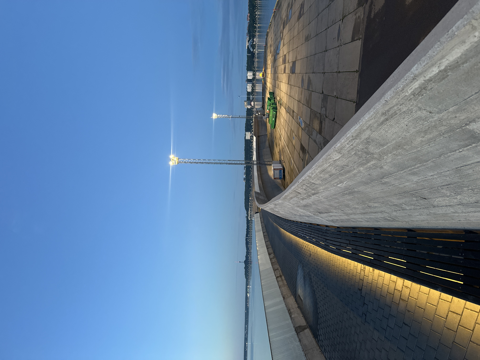 The image shows a pier in Tallinn, Estonia at dusk. The sky is a deep blue, transitioning to lighter shades near the horizon. A long, curving concrete barrier dominates the foreground, with a paved walkway and benches on the left, illuminated by warm yellow lights. The right side of the pier is a large, open, paved area with square concrete slabs. Two tall light poles with bright lights stand on the pier, illuminating the scene. In the distance, across the water, the cityscape of Tallinn is visible, with buildings, lights, and a prominent radio tower. There are no people or animals in the image. The atmosphere is calm and serene, with the lights reflecting on the wet pavement, suggesting recent rain. Green industrial equipment is visible on the pier, and there is a yellow object in the distance that might be a vehicle.