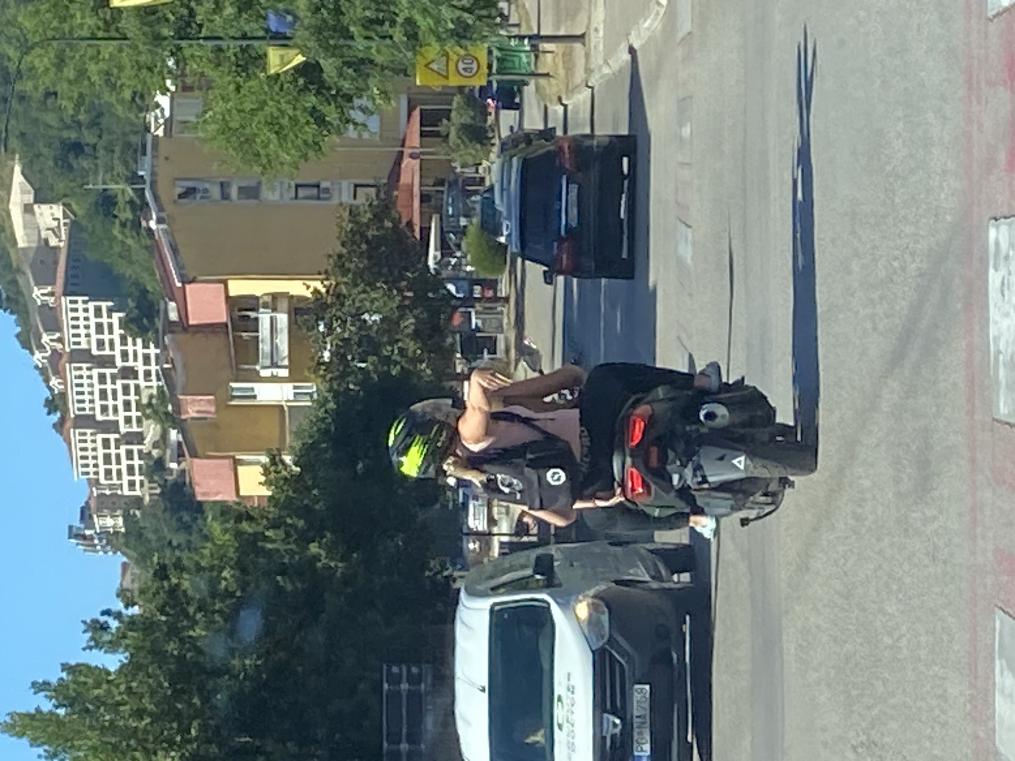 The image shows a street scene in Budva, Montenegro, on a sunny day. Two people are riding a scooter. The person in the front is wearing a pink tank top, black pants, and a black backpack with a white logo. They have blonde hair and are wearing a helmet with green and black accents. The person in the back is wearing black pants and a helmet. They are holding onto the person in the front. A white van is to the left of the scooter, and a dark SUV is to the right. In the background, there are buildings on a hillside, trees, and a street sign with a 40 km/h speed limit. The road surface appears to be asphalt, with a pedestrian crossing marked by white and red lines in the foreground. The overall impression is a typical urban street scene during daylight hours.