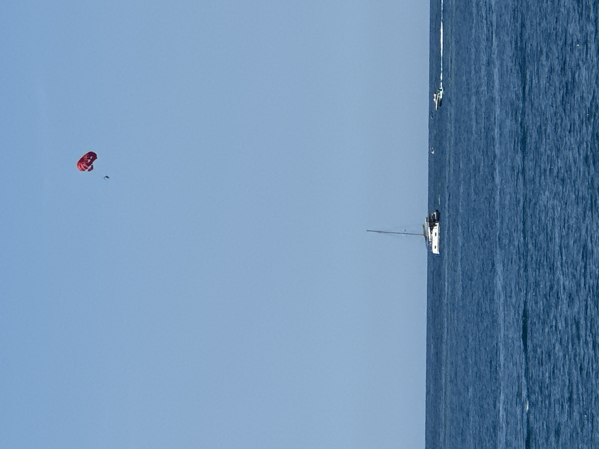 This vertical full shot captures a clear day over the Adriatic Sea near Budva, Montenegro. The upper two-thirds of the frame consists of an unobstructed light blue sky. The lower third features dark blue ocean water with subtle surface ripples.
In the upper right quadrant of the sky, a red parasail is visible, with a single individual, a dark silhouette, suspended directly beneath it. The parasail appears partially inflated or in a state of descent/ascent.
On the water, a white catamaran with a mast is positioned slightly left of the vertical center, appearing to be sailing or anchored. Further to the right and slightly beyond the catamaran, a white motorboat is in motion, generating a distinct white wake behind it. Additional small, indistinct watercraft are barely perceptible near the horizon line in the far distance, positioned between the catamaran and the moving motorboat. No land is visible in the frame.