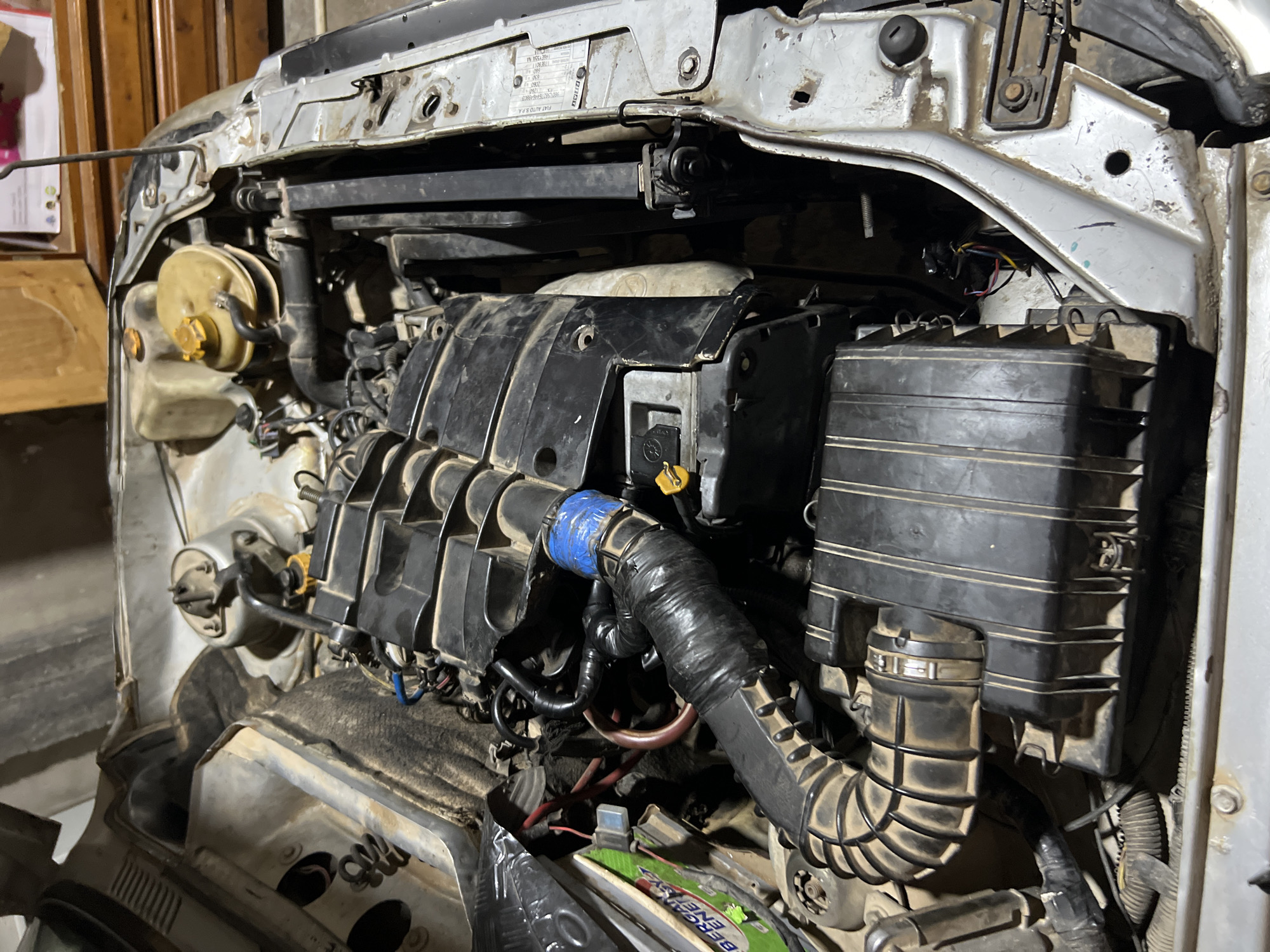 This is a forensic, matter-of-fact description of an engine bay, potentially in Sidi Chahmi, Algeria. The image shows a close-up view of a car engine, with various components visible. The engine appears to be dirty and dusty, suggesting it may be old or has been exposed to the elements. Several hoses, wires, and plastic parts are present, along with what appears to be a battery. The overall condition of the engine suggests it might be undergoing maintenance or repair. There are no people or specific actions depicted in the image, focusing solely on the mechanical components of the vehicle. The environment appears to be an indoor setting, possibly a garage or workshop, with wooden structures and boxes visible in the background.