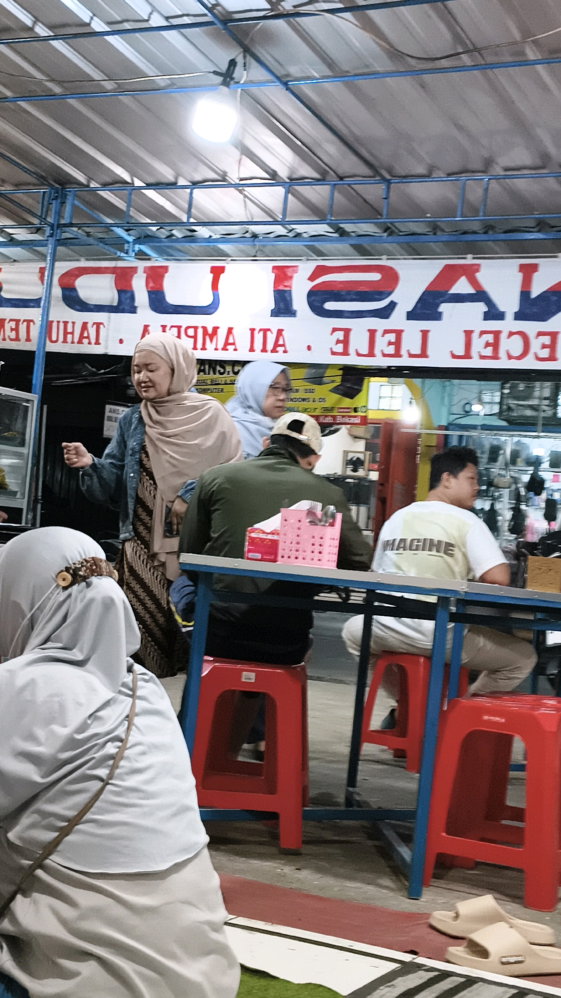 In Karangsatria, Indonesia, a group of individuals is gathered at a food stall under a corrugated metal roof supported by blue metal framing. Two women, one in a light brown hijab and denim jacket, and another in a light blue hijab, are standing near the stall. The woman in the brown hijab is gesturing with her left hand. In front of them, at a blue metal table with red plastic stools, three people are seated. One person, facing away from the camera, wears a dark green jacket and a beige cap. Another, also facing away, is in a white t-shirt with "IMAGINE" printed on the back. The third person, partially visible, is wearing a white shirt and beige pants. On the table, a pink plastic container holds cutlery. A banner with large, red and blue lettering is displayed behind the stall. The ground is covered in a mix of red and green mats. A pair of beige sandals rests on the mats near the red stools.