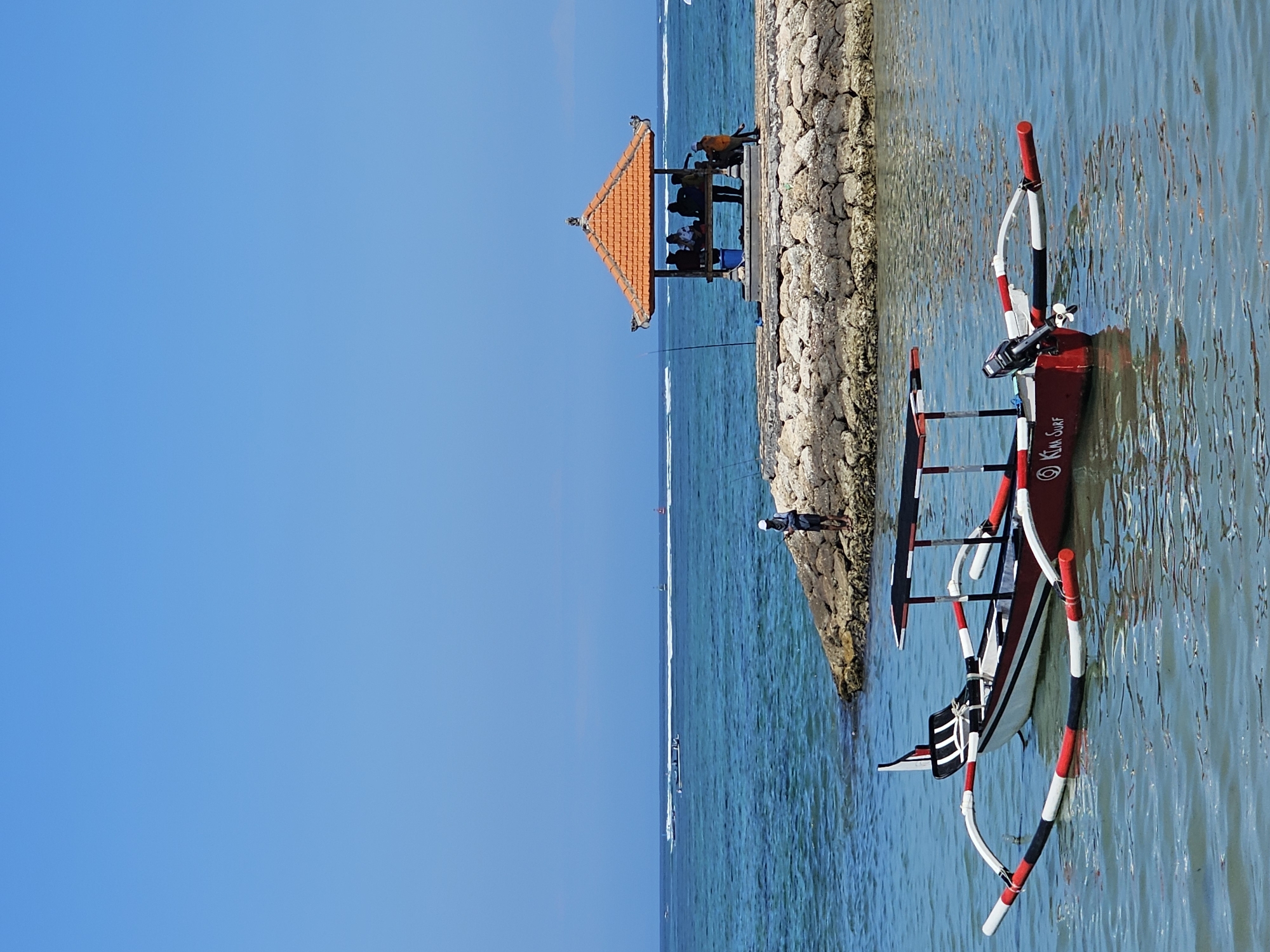 A medium shot captures a serene coastal scene at Serangan Beach in Denpasar, Indonesia, on a clear, sunny day. In the foreground, a traditional Indonesian outrigger boat, painted red with white and black accents, rests on the calm, blue water. The boat features a simple canopy for shade and an outboard motor mounted at the stern.
In the mid-ground, a man stands on a stone jetty, fishing with a rod. Beyond him, a small, orange-roofed gazebo offers shelter to a group of people. The gazebo is situated on a rocky breakwater, extending into the turquoise ocean. The water exhibits a vibrant blue hue, with gentle waves and white wakes from distant boats. The horizon is marked by the clear, unbroken line of the sea and sky. The overall atmosphere is peaceful and tranquil, indicative of a typical day at this popular beach destination.