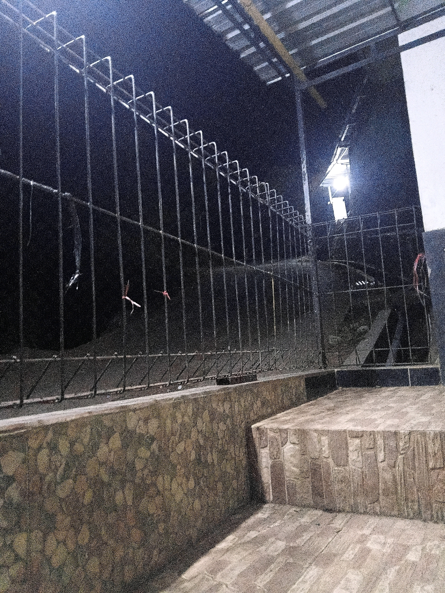 The image depicts a nighttime scene in Sinduaji, Indonesia, focused on a metal fence that runs horizontally across the frame. The fence is constructed of vertical bars and has a decorative zigzag pattern along its top. Behind the fence, the environment is dark and appears to be an outdoor area with a paved surface. A bright light illuminates a portion of the scene from the upper right. The lower part of the image shows a textured wall and a step made of stone-like material. There are faint traces of smoke or debris visible beyond the fence.