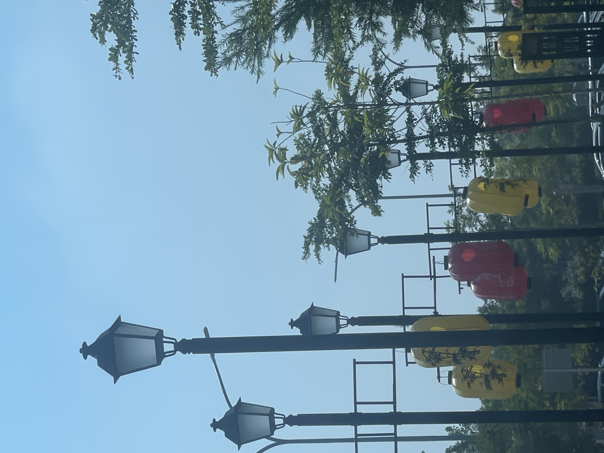 The image depicts a street scene under a clear blue sky. Several black lampposts are visible, some with decorative lanterns hanging from them. The lanterns are of two colors: yellow, with black painted designs that appear to be bamboo, and red, with some text visible on one of them, though it is difficult to read clearly. Green trees and foliage are present, interspersed with the lampposts and lanterns, suggesting an outdoor urban environment. The scene appears to be a public space, possibly a park or a pedestrian walkway, in East Jakarta, Indonesia, given the presence of these decorative elements common in some parts of the city. There are no people or vehicles clearly visible in the immediate foreground.