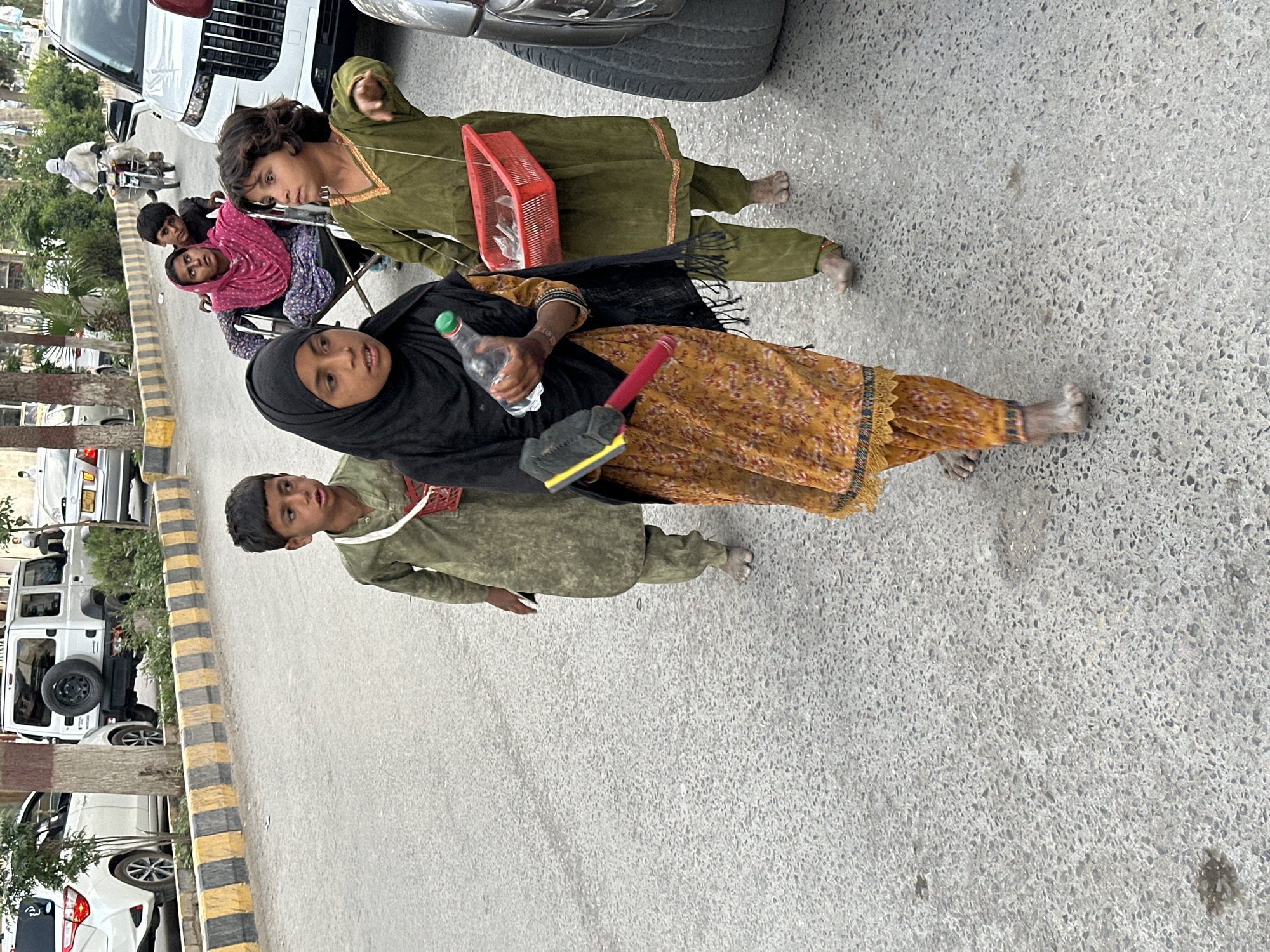 The image shows a group of people on a street. A woman in a black hijab and yellow dress is holding a water bottle and a red object with a black sponge. A boy in a green outfit is standing beside her. Another child is holding a red basket. Other individuals are seated in the background. The location is Quetta City Tehsil, Pakistan. Cars are parked along the side of the road.