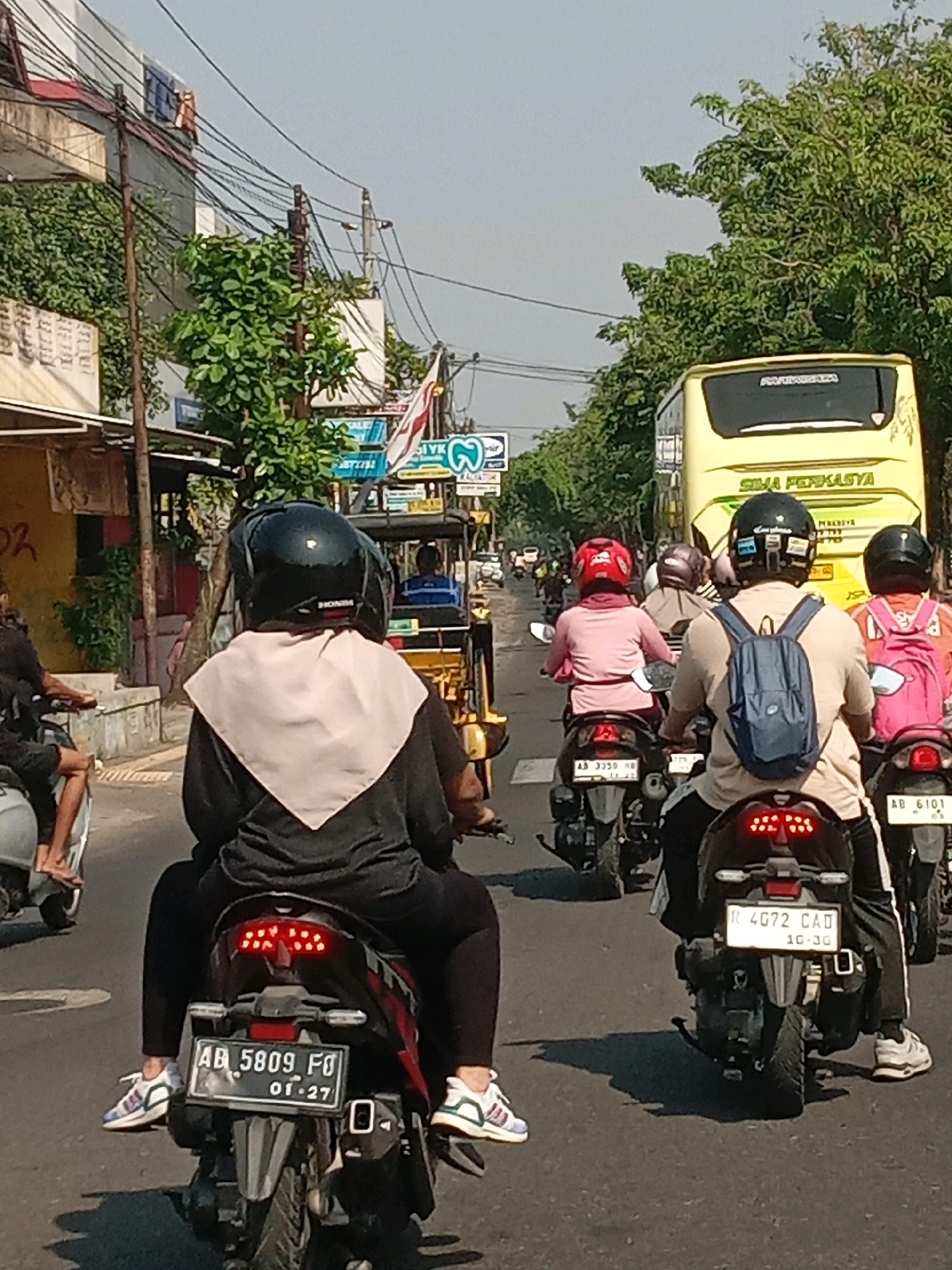 The image depicts a street scene in the city of Yogyakarta, Indonesia, likely on a sunny day given the clear sky and bright lighting. The foreground is dominated by a black motorcycle with a license plate reading "AB 5809 FO" and "01-27". Two people are seated on this motorcycle, the rider wearing a black helmet and the passenger wearing a light-colored headscarf and a black garment. Their feet are visible, with the passenger wearing white sneakers with colored accents.
To the right of the foreground motorcycle, another black motorcycle is visible with a license plate "R 4G72 CAD" and "10-30". Two individuals are on this motorcycle as well. The passenger is wearing a pink shirt and a red helmet, while the rider is wearing a light-brown shirt and a dark helmet with a blue backpack.
Further ahead, a yellow bus with the text "SIMA PERKASYA" on its rear is traveling in the same direction. Several other motorcycles and a three-wheeled vehicle are also present on the road, indicating moderate traffic. The roadside features buildings with advertisements and signage, as well as lush green trees. Overhead, a complex network of electrical wires crisscrosses the sky, a common sight in urban Indonesian environments. The overall impression is a bustling street in a typical Indonesian city.