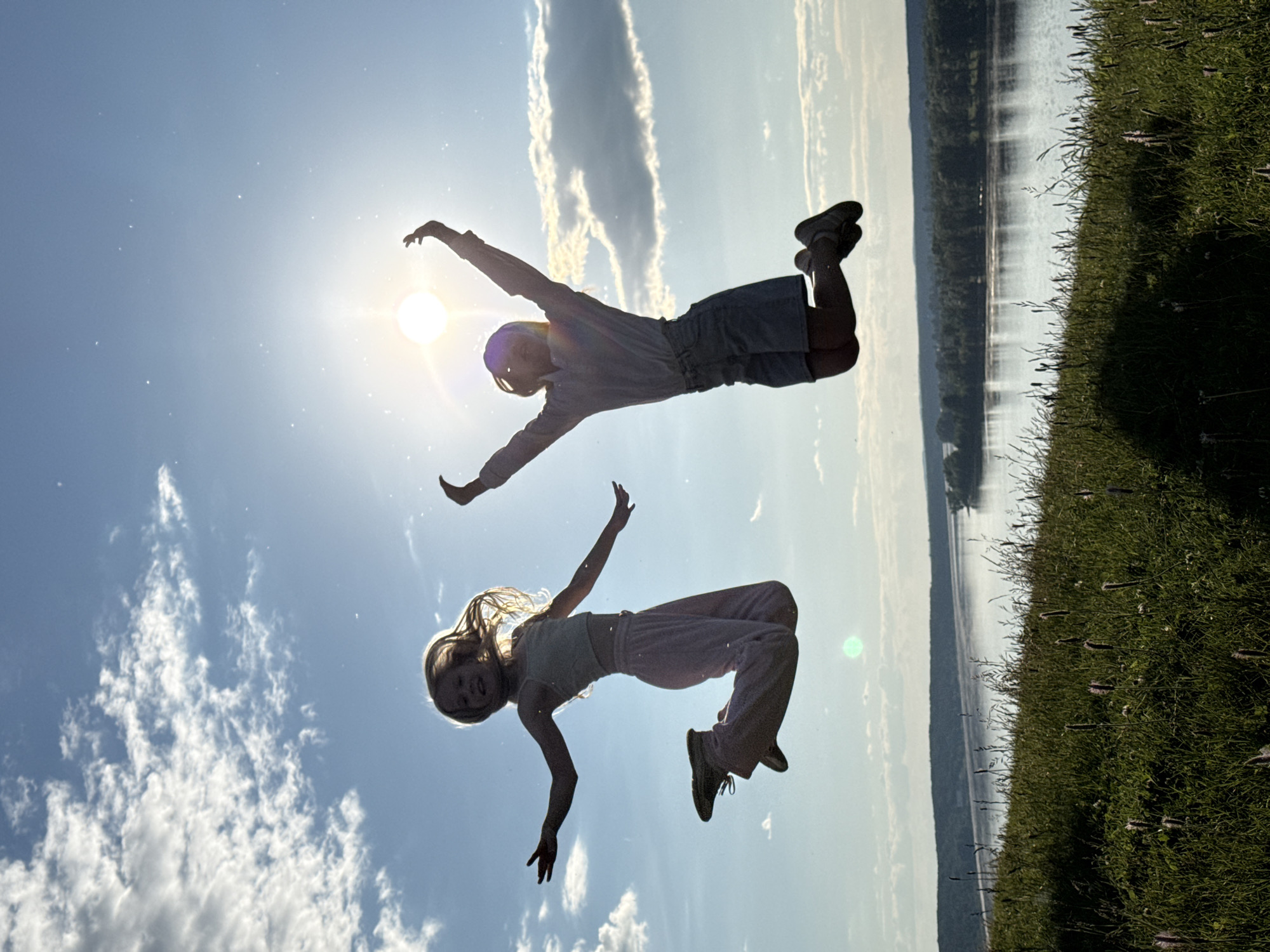 This image captures two young girls in mid-air against a bright sky, indicative of a jump, on a sunny day in the city of Ольховка, Russia.
The scene is set outdoors, with a wide river and a distant treeline visible in the background, bordered by a grassy hill in the foreground. The sun, a prominent bright circle with lens flare, is positioned in the upper right quadrant of the sky, backlighting the subjects. The sky is clear blue with scattered white cumulus clouds, particularly on the left side. Small light particles are visible in the air, illuminated by the sun.
Two girls are the primary subjects, both silhouetted but with visible details. The girl on the left has blond hair, illuminated by the backlighting, and appears to be smiling. She is wearing a light-colored crop top and light-colored long pants, with her arms spread wide and her legs bent with knees drawn up. Her body is angled slightly to the right. The girl on the right is also in mid-air, with her arms raised high above her head in a celebratory gesture and her legs bent with knees drawn up. She is wearing a light-colored long-sleeved top and light-colored shorts. Her face is mostly obscured by shadow due to the strong backlighting. Both girls' poses suggest an active jump.
The foreground consists of green grass with various wild plants and darker shadow patches, providing a textured surface leading down towards the river. The river itself reflects the sky, appearing calm, with a dense forest strip along the opposite bank, and hazy hills visible further in the distance.
