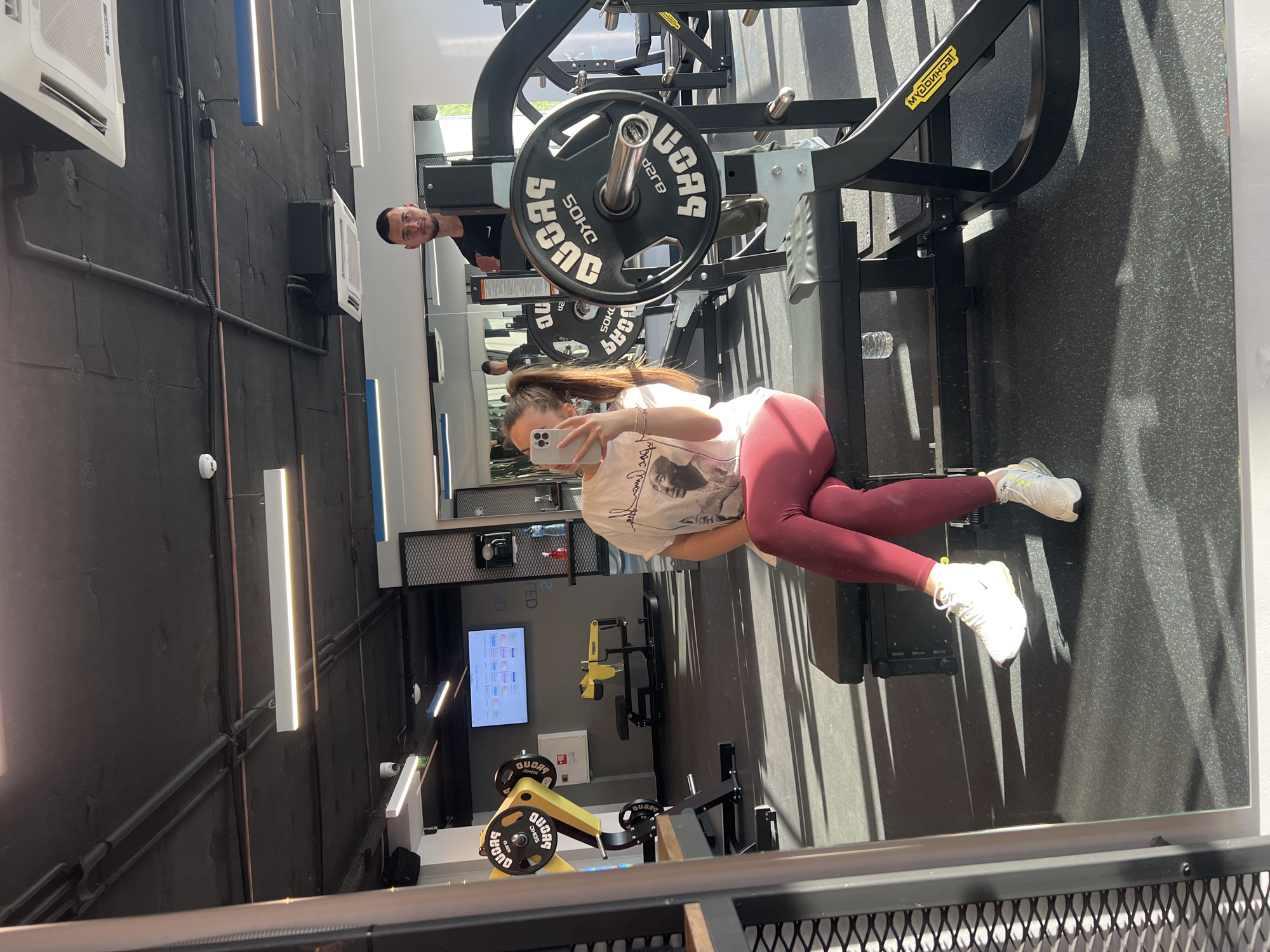 This image captures a mirror selfie taken inside a modern gym in Plovdiv, Bulgaria, during the daytime, evidenced by strong shadows cast by sunlight.
The primary subject is a woman with long brown hair in a ponytail, wearing a white graphic t-shirt, burgundy leggings, and white sneakers. She is seated on a gym bench or machine, holding a light-colored smartphone to take the photo, obscuring her face. Reflected behind her, a man with short dark hair and a beard looks towards the camera with a neutral expression.
The gym features a sleek, industrial design with black walls, exposed pipes, and rectangular LED light fixtures, some emitting white light, others a blue hue. Several white air conditioning units are mounted on the walls. Various gym equipment is visible, including a squat rack or similar machine with a large black "50KG" weight plate, branded "QUORA" and "TECHNOGYM." Another yellow-framed machine also bears the "QUORA" brand. A water bottle rests on the floor near the woman. In the background, a digital screen displays information, and security cameras are mounted on the walls. Due to the mirror reflection, all visible text and details appear reversed.