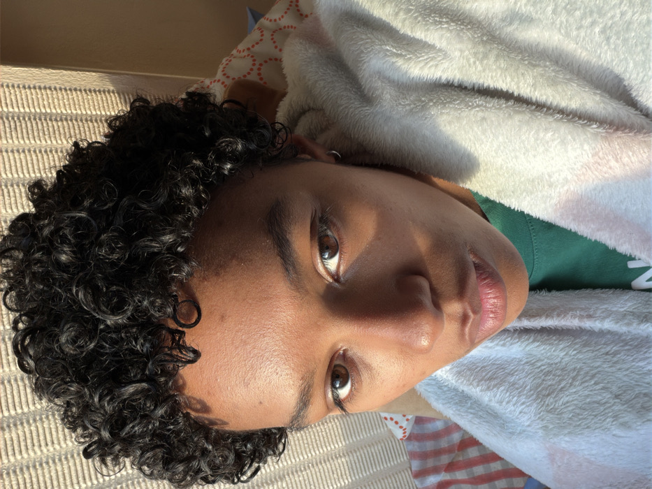 The scene, located in Champs-sur-Marne, France, presents a close-up, eye-level indoor portrait of an individual. The subject has dark skin, short, dark, tightly curled hair, and dark brown eyes, maintaining direct eye contact with the viewer. A small silver hoop earring is present in the subject's left ear. The subject is wearing a green shirt and partially enveloped in a light-colored, plush textile, consistent with a blanket or robe. Illumination originates from the upper right, creating pronounced highlights on the right side of the face and casting shadows across the left side and forehead. The background elements are blurred: the upper left shows a light-toned, vertically textured surface; the upper right, a solid light brown wall. Fabric patterns are visible below, including red and white stripes on the left and faded orange circular motifs on a lighter ground on the right.