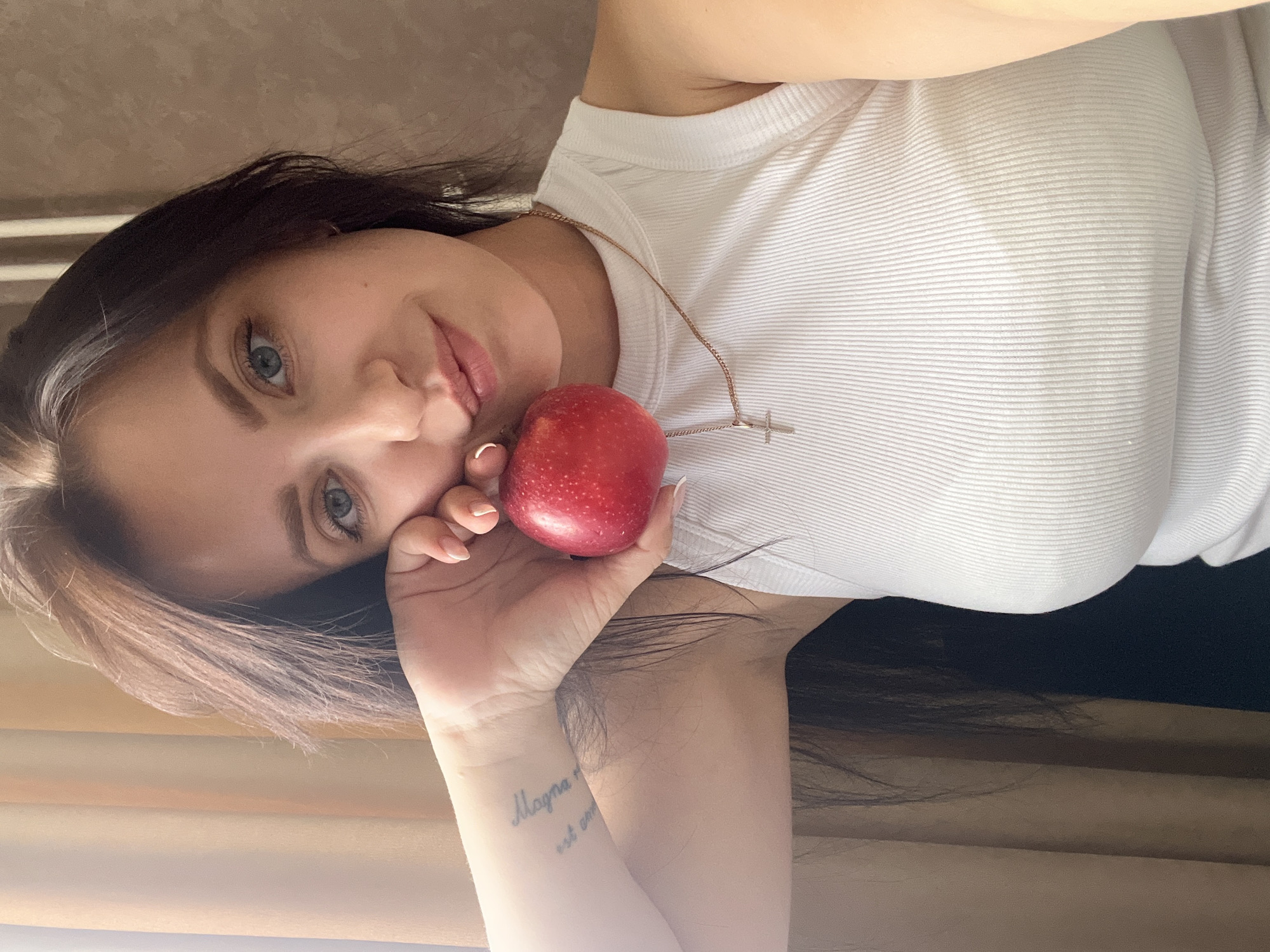 A young woman with dark hair and blue eyes is holding a red apple in her hand. She is wearing a white ribbed tank top and a thin gold chain with a cross pendant. A tattoo is visible on her left forearm. The background consists of curtains and a wall. The image appears to be a selfie taken indoors. There is no visible indication of the specific location within Марковский, Russia.