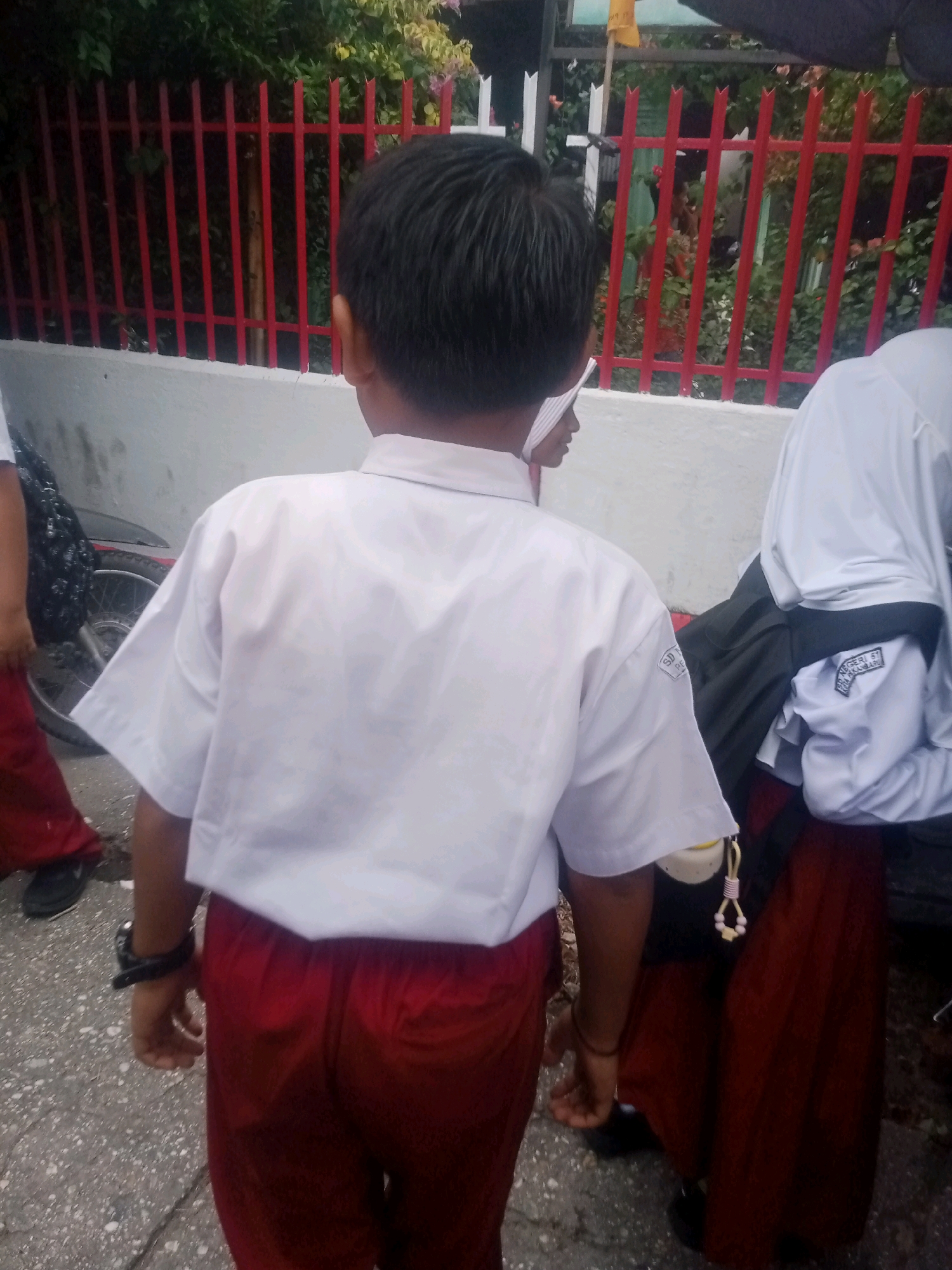The image shows a group of young people, likely students, in what appears to be an outdoor area near a school in Pekanbaru, Indonesia. The central figure, seen from behind, is wearing a white short-sleeved shirt and red pants, with a black watch on their left wrist. To the right, another individual is wearing a white hijab and a dark skirt, with a patch on their white shirt that reads "SD NEGERI 51 PEKANBARU". A third person, partially visible on the left, is wearing red pants. In the background, a red metal fence with vertical bars and some greenery can be observed, along with a light-colored wall. The ground is paved with asphalt and small stones. The overall scene suggests a casual outdoor gathering or movement of students.