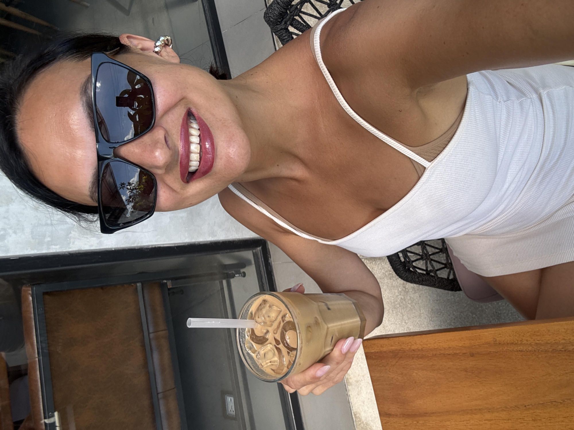A woman with dark hair and sunglasses is photographed at a slight angle, holding a glass of iced coffee with a straw. She is wearing a white tank top dress. Her manicured nails are visible as she holds the drink. The reflection in her sunglasses shows greenery and a building exterior. The background includes a glass-encased area with a brown cushioned seat and a rattan chair. The location is North Kuta, Indonesia.