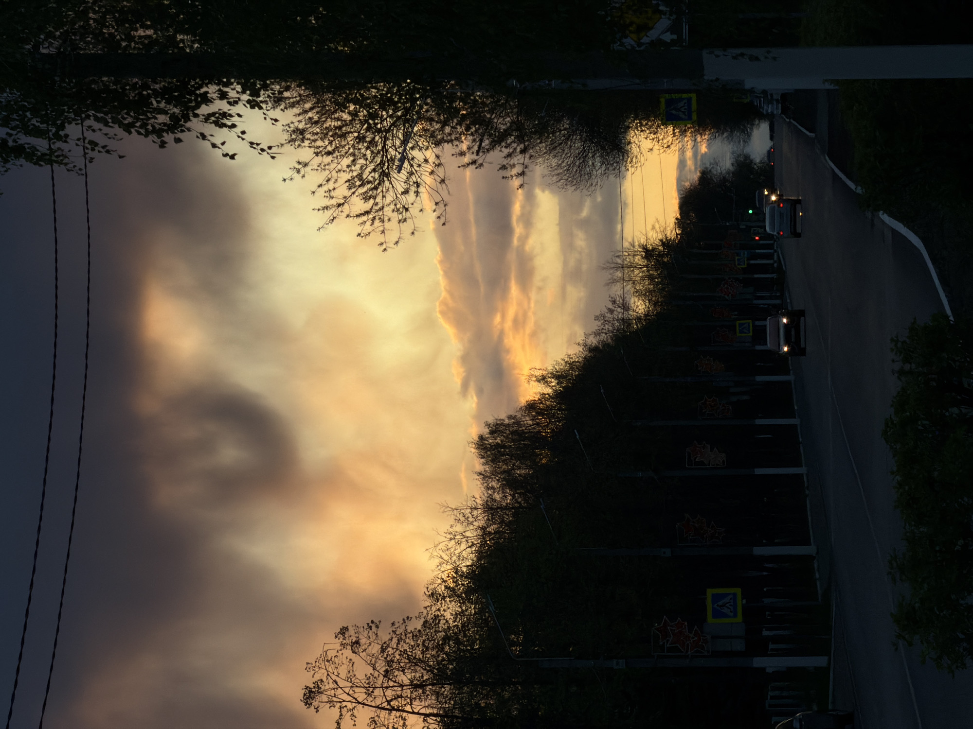The image shows a street in Chaikovsky, Russia at sunset. The sky is filled with orange and grey clouds. The street is lined with trees and streetlights, some of which are adorned with star-shaped decorations. Several vehicles with headlights on are visible on the road. Pedestrian crossing signs are seen along the road. Electrical wires are visible on the left side of the image.