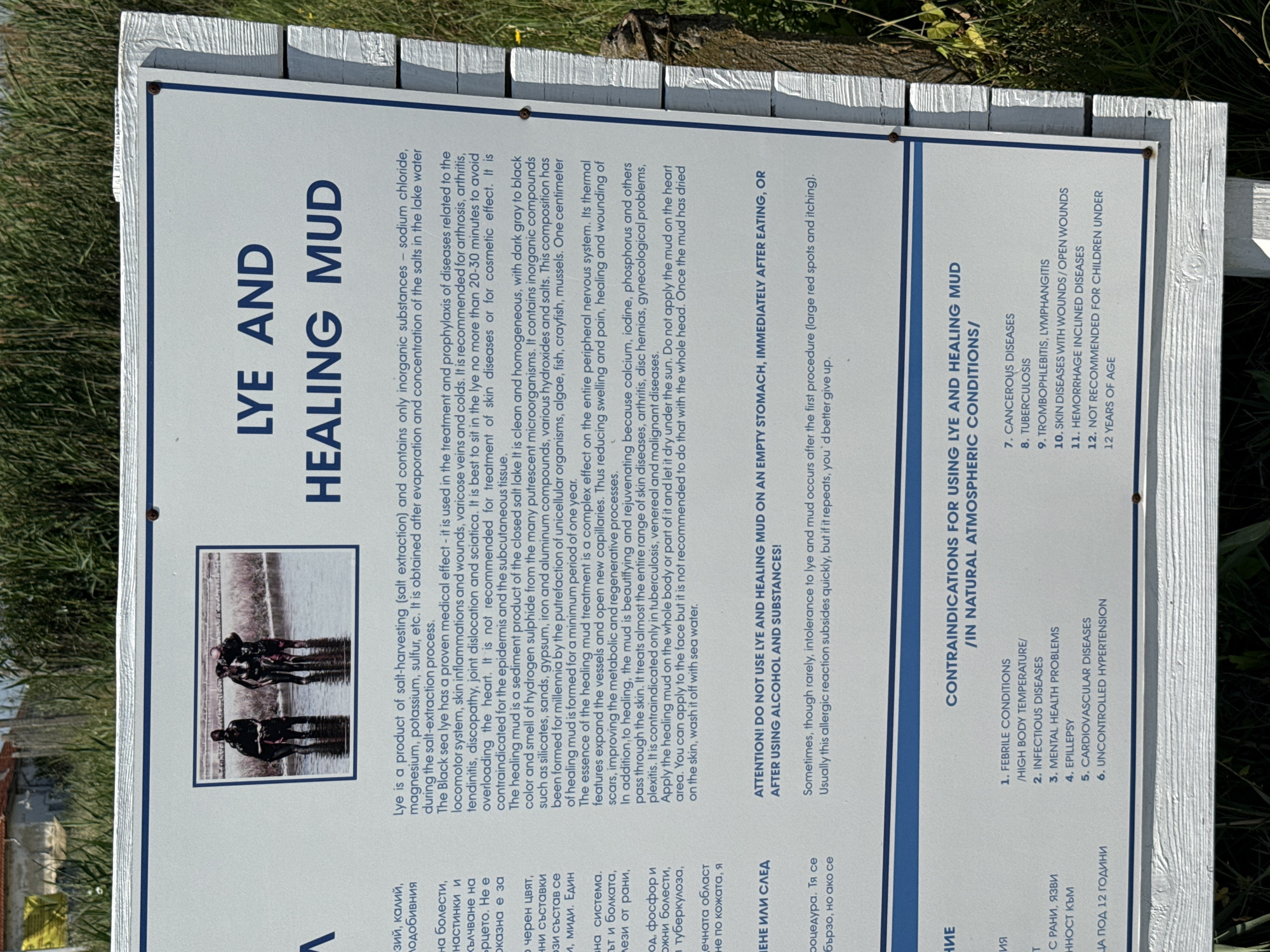 The image shows a sign in Bulgarian and English about "LYE AND HEALING MUD" located at the Burgas Saltworks in Bulgaria. The sign has a large, central photograph of four individuals, two men and two women, wading in shallow water, appearing to be harvesting or working with mud. The text on the sign describes the properties and uses of lye and healing mud, its medical effects, contraindications, and recommended procedures. It mentions that the lye is a product of salt-harvesting and contains inorganic substances. The healing mud is described as a sediment product of closed salt lake color and is formed for millennia. The sign also warns against using lye and healing mud on an empty stomach or after consuming alcohol. In the lower section of the sign, a list of contraindications for using lye and healing mud in natural atmospheric conditions is provided, including febrile conditions, infectious diseases, mental health problems, epilepsy, cardiovascular diseases, cancerous diseases, tuberculosis, thrombophlebitis, lymphangitis, skin diseases with wounds, hemorrhage inclined diseases, and a recommendation not to use for children under 12 years of age. The environment depicted is outdoors, with the sign set against a backdrop of vegetation. The sign itself is white with blue lettering and borders, mounted on a white wooden post.