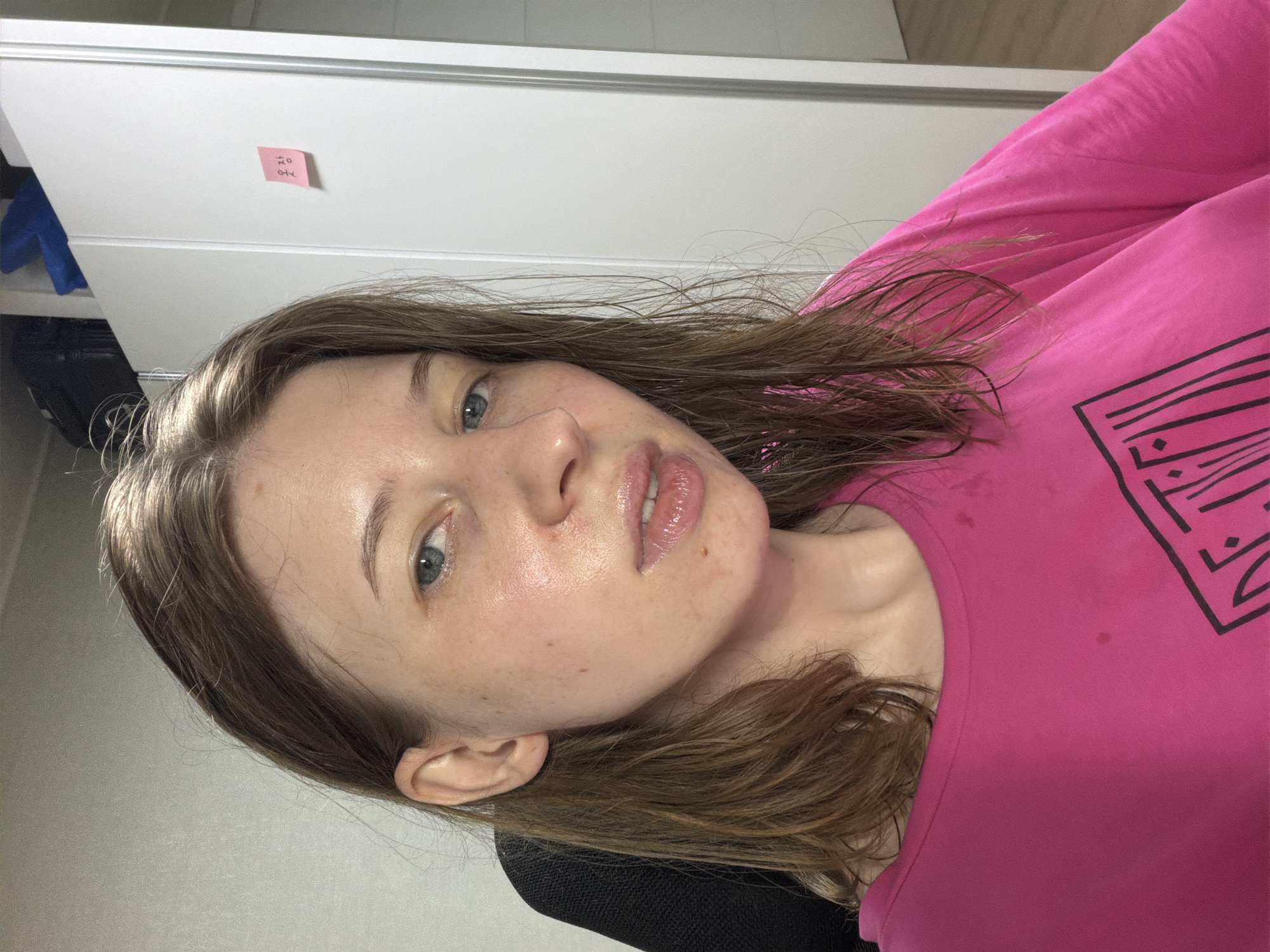 A close-up, over-the-shoulder selfie shows a young woman with light brown hair that is wet and extends past her shoulders. She is wearing a bright pink t-shirt. Her face is illuminated, highlighting the sheen of her skin and her blue eyes. She has some freckles and a small mole on her cheek. Her lips are glossy, and her mouth is slightly open. She is positioned in front of a tall, white closet. On the closet door, a small pink sticky note is affixed, bearing Korean characters that read "옷장" (otjang), meaning "closet." The background also shows a black office chair and a glimpse of a wooden floor. The lighting suggests an indoor environment, possibly a bedroom or living space, within a residence in Dongducheon-si, South Korea.